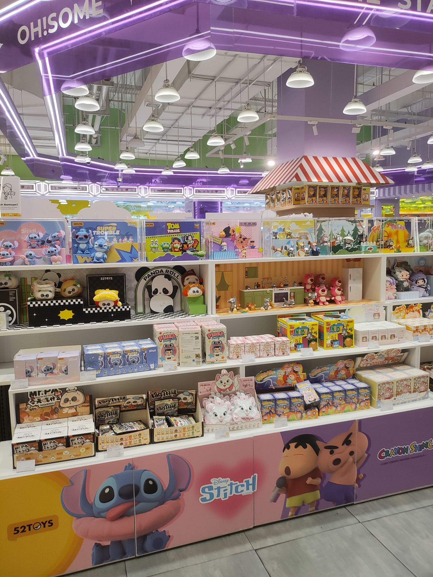 The image shows a retail store interior in Lambangjaya, Indonesia, likely a toy store, based on the abundance of merchandise. The store features brightly colored displays and shelves stocked with various toys, including plush characters, action figures, and collectible items. Prominently displayed are merchandise featuring Disney's Stitch, characters from Toy Story, and possibly Crayon Shin-chan, indicated by the signage. The overall environment is clean, well-lit with overhead fluorescent lights, and organized to showcase the products. The ceiling has a unique purple lighting design with the text "OH!SOME" visible.