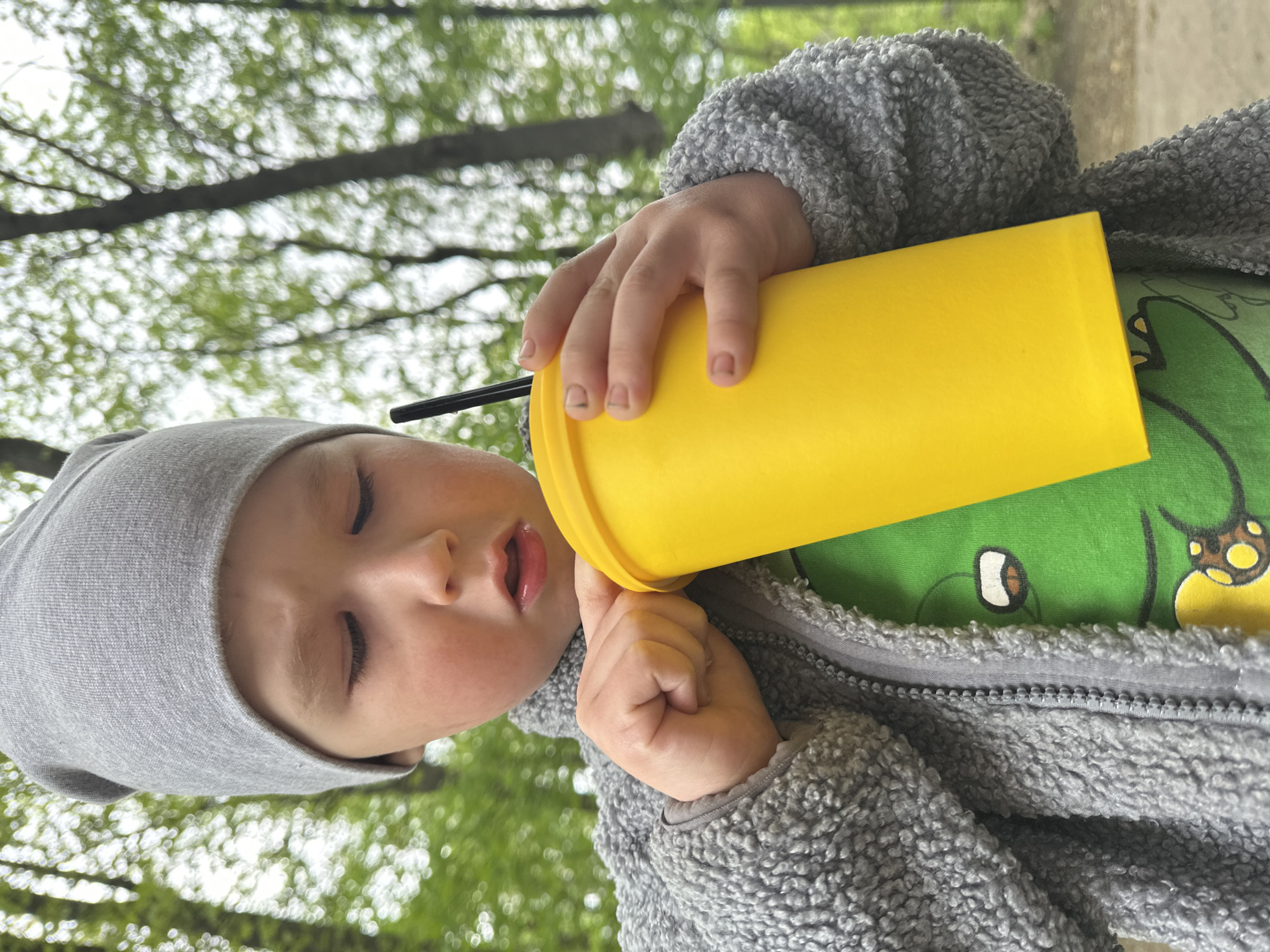 A young child with light skin, wearing a grey beanie and a green shirt with a dinosaur graphic under a grey fleece jacket, is holding a yellow plastic cup with a black straw to their mouth. The child's eyes are mostly closed. The background shows blurred foliage and tree branches, suggesting an outdoor setting. The photo appears to have been taken outside, perhaps in a park or garden. There is no indication of the location being in Nizhny Novgorod, Russia or anywhere else in particular.