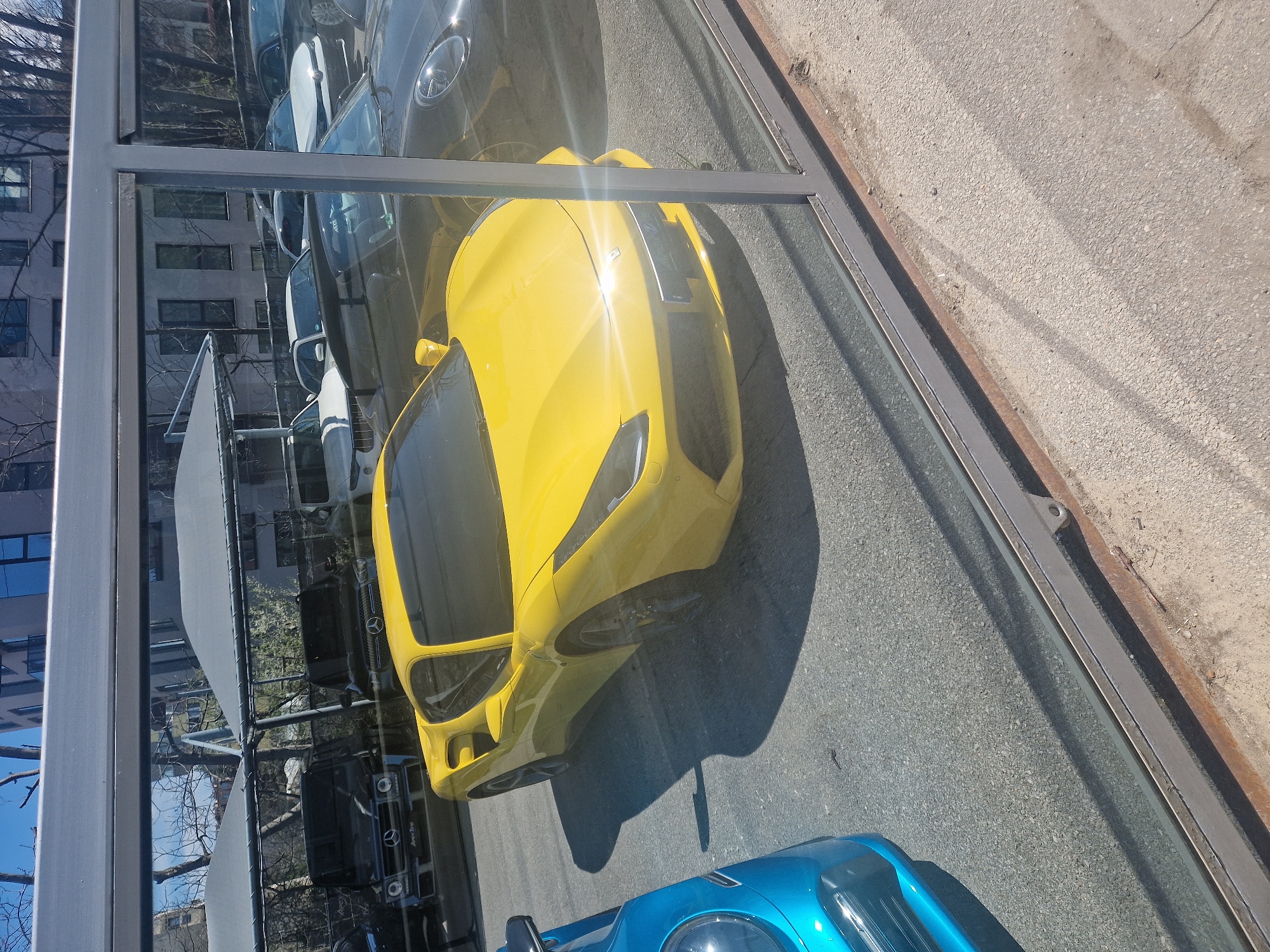 The image captures a street scene in Unknown, Unknown, viewed through a glass window. Prominently featured are several parked cars, including a bright yellow sports car dominating the foreground and a blue car partially visible at the bottom of the frame. A black vehicle is parked nearby. The reflection in the glass reveals a building, bare trees, and other parked cars. A metal frame encases the glass. The ground is asphalt.
