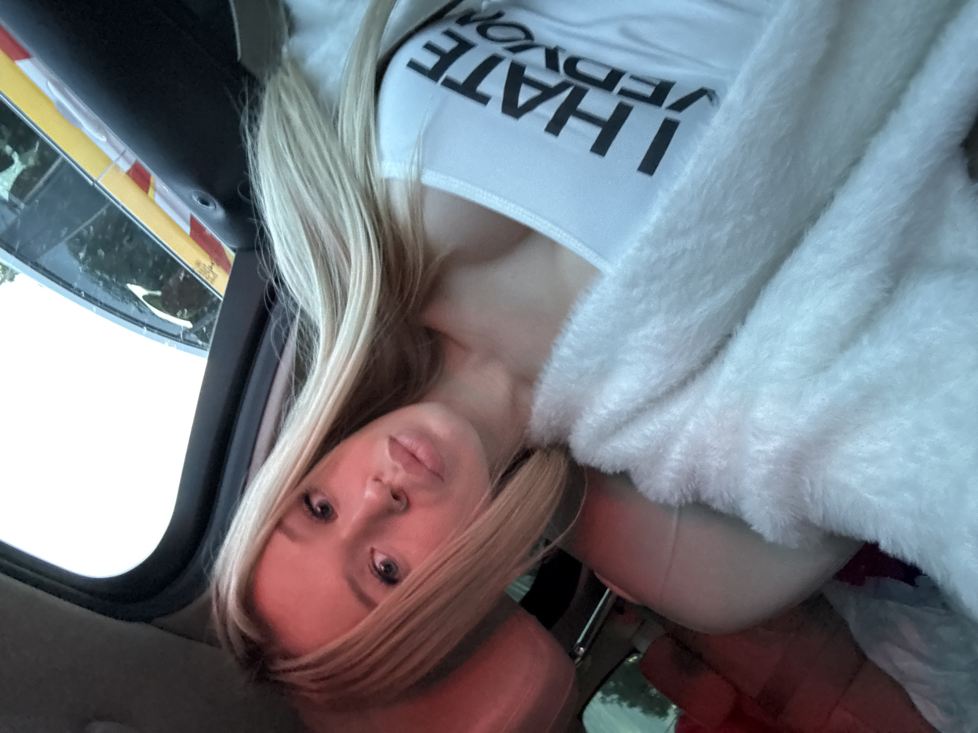 The image is a close-up, vertically oriented selfie taken from inside a vehicle. The main subject is a young woman with blonde hair, wearing a white tank top with bold black lettering that reads "I HATE VEGANS". She is also wearing a fluffy white garment, possibly a jacket or shawl, draped over her shoulders and chest. The woman's face is partially visible, tilted towards the camera, with her eyes looking directly at the lens. Her expression appears neutral or perhaps slightly bored.
The setting appears to be the interior of a car. Through the window on the left, a blurred view of what looks like a yellow and red striped barrier or gate is visible, suggesting the car might be at a toll booth or an entry point. The lighting suggests it could be daytime, though the overcast sky outside contributes to a somewhat dim overall lighting. The colors in the image are muted, with the white of her clothing and the fluffy material contrasting with the darker interior of the car and the soft pinkish hue on the woman's face, possibly from ambient light.
The image does not convey a specific event or activity beyond the act of taking a selfie. There are no explicit location cues tying it to Saint Petersburg, Russia, beyond the provided context.