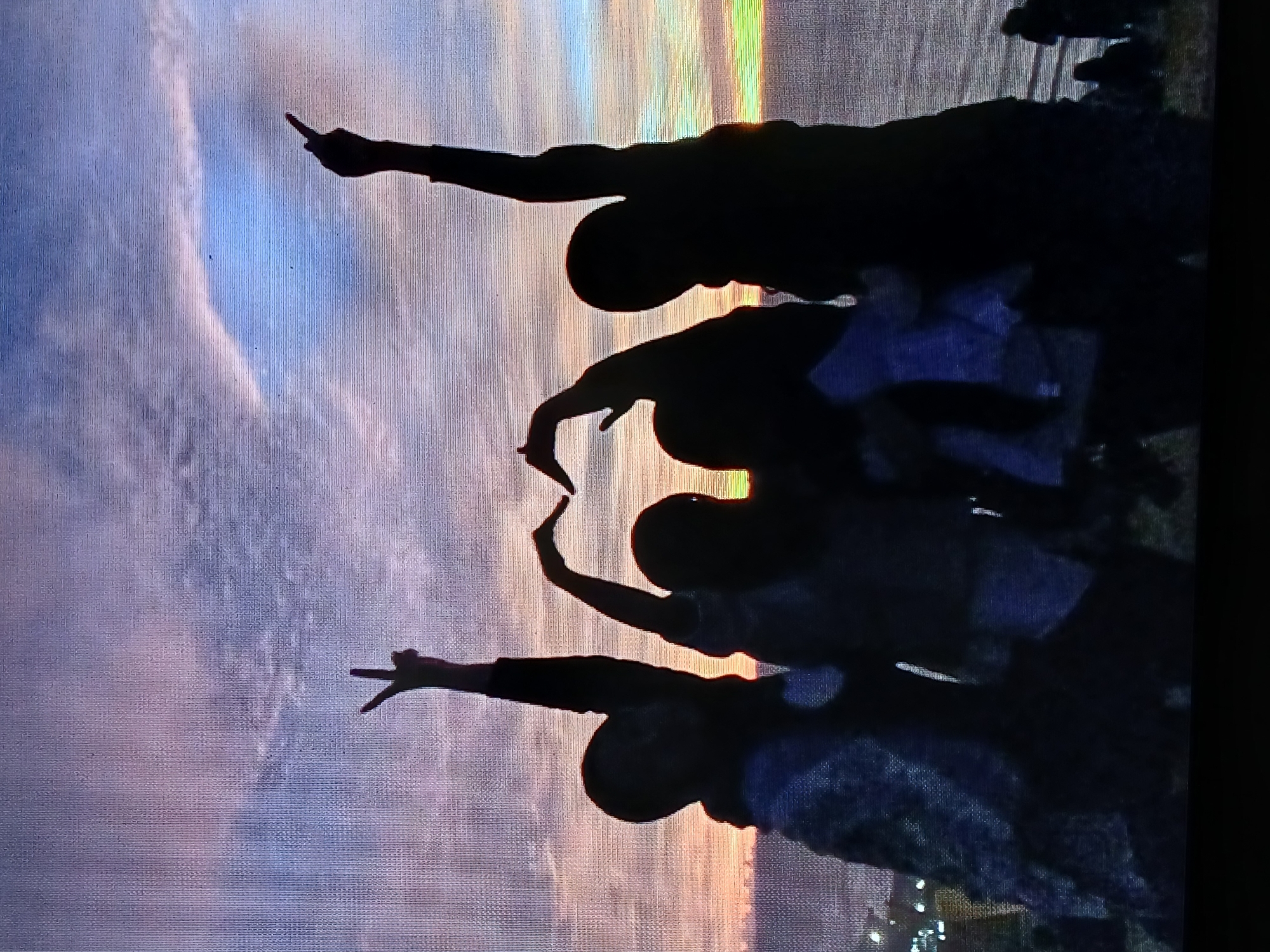 The image depicts a group of people silhouetted against a sunset sky, with at least three individuals visible making hand gestures. One person points a finger upward, another forms a heart shape with their hands, and a third makes a peace sign. The background features a gradient of colors transitioning from purple and blue to orange and yellow, suggesting a sunset. A landmass or coastline is visible at the right side of the image, with some faint lights visible, possibly indicating the urban area of Juan Griego, Venezuela.