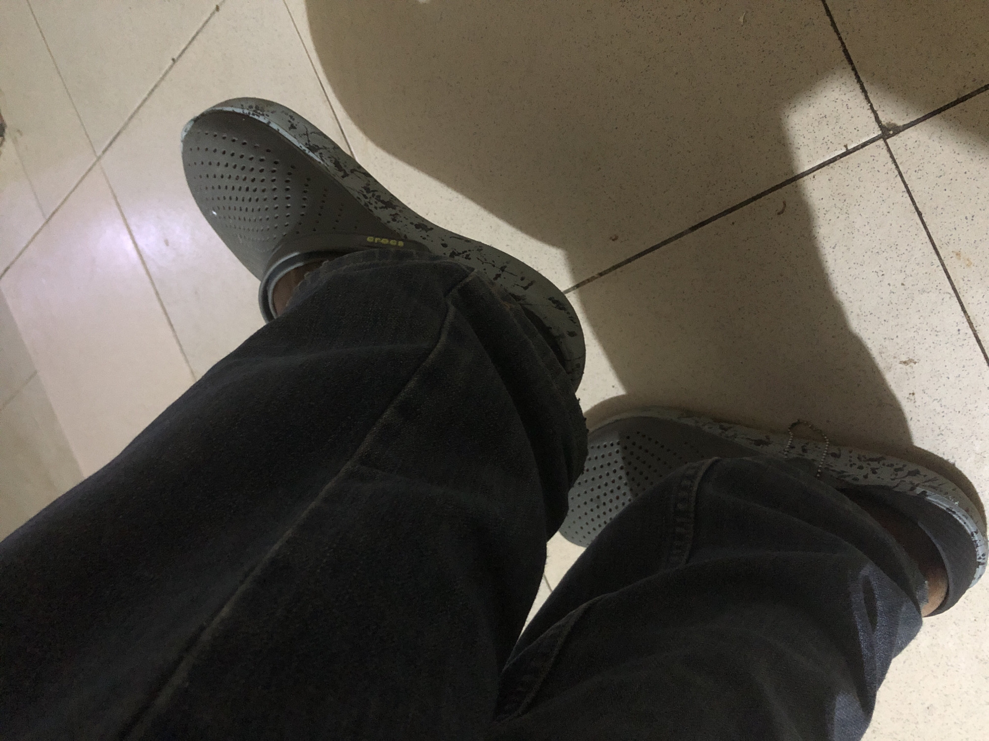 In Kabong, Nigeria, an individual is seated with their legs crossed, wearing dark wash denim jeans and a pair of grey Crocs clogs. The clogs have a perforated design on the upper and a speckled pattern on the sole, with visible wear and tear on the edges. The person's right foot is resting on the left. The flooring is tiled with a light-colored, speckled pattern, and a shadow is cast on the tiles to the right of the subjects. The lighting suggests an indoor environment.