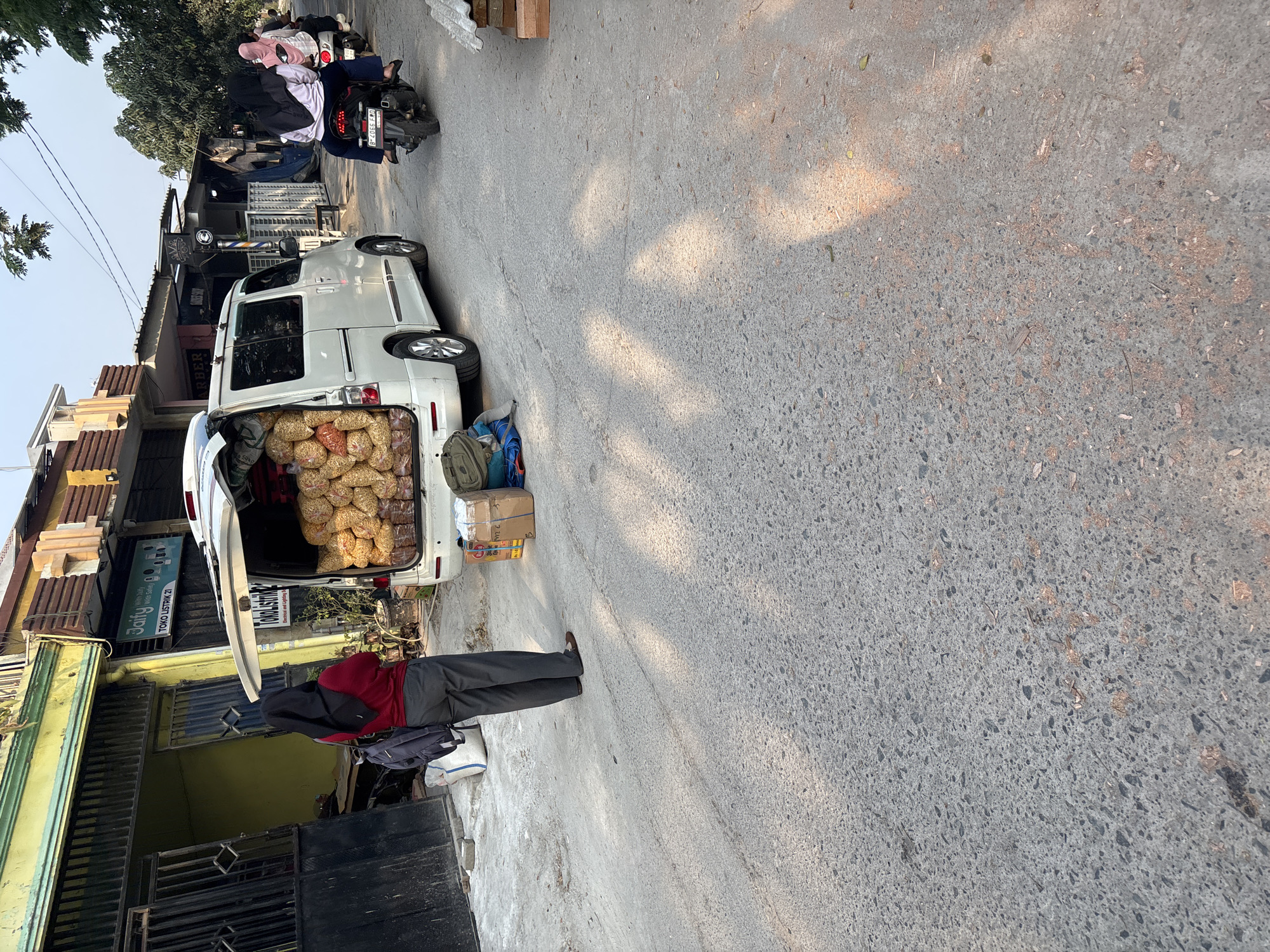 The image shows a street scene in Telagamurni, Indonesia. In the foreground, a white van is parked on a paved road, its rear doors open to reveal a large quantity of bagged snacks. A person wearing a red hijab and dark pants stands near the van, facing away from the camera, with a backpack on their shoulders. Several bags and boxes are scattered around the back of the van. In the background, two people on a motorcycle are riding away from the camera. The buildings lining the street appear to be shops and businesses. The overall atmosphere suggests a commercial or delivery setting in a residential or mixed-use area.