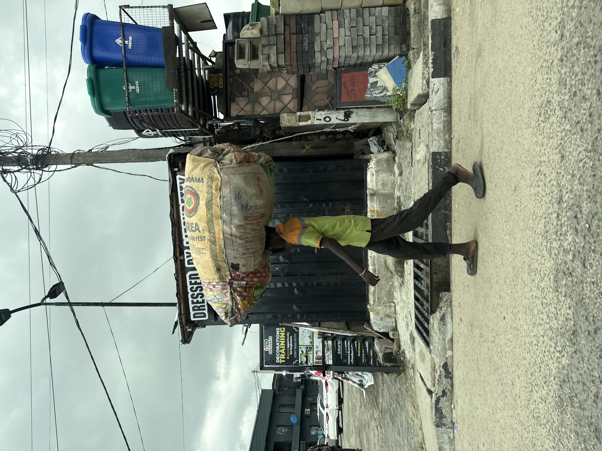 A man carries a large sack on his head while walking along a concrete sidewalk in Ado, Nigeria. The sack is labeled "DRESSED." Behind him, there is a building with a sign advertising "DECORATIONS TRAINING." In the background, there are trash bins, bricks, and electrical wires. The sidewalk is adjacent to a road.