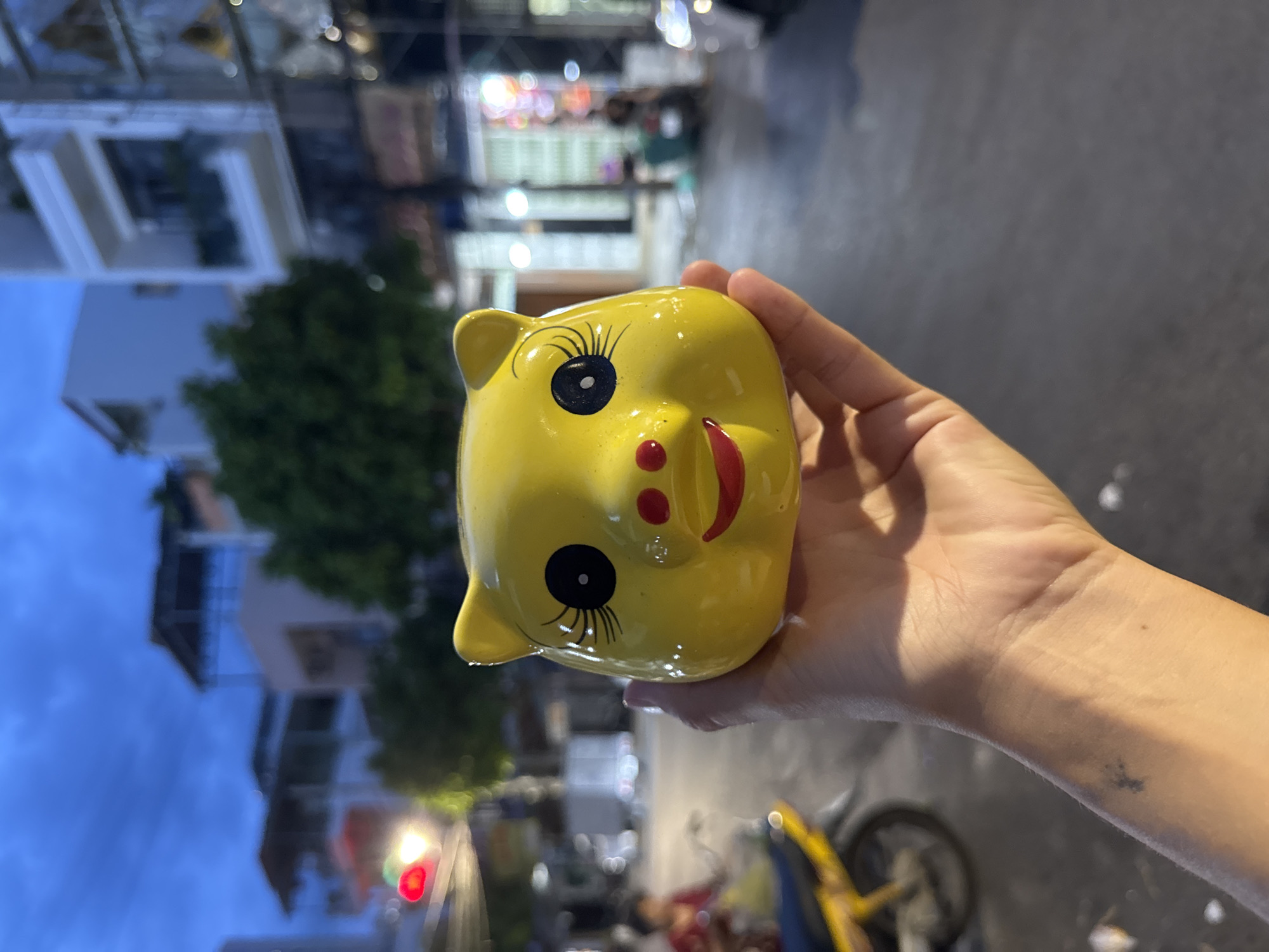A close-up shot shows a person's hand holding a bright yellow, ceramic piggy bank shaped like a pig's head. The piggy bank has large, round black eyes with eyelashes, two small red dots for nostrils, and a red smile. The background is blurred and depicts a street scene in Nha Trang, Vietnam, at dusk. Buildings with multiple stories line the street, and a traffic light with red and yellow lights is visible on the left. A yellow motorcycle is parked in the middle ground, and a person is partially visible on it. The overall atmosphere suggests an urban environment at the end of the day.