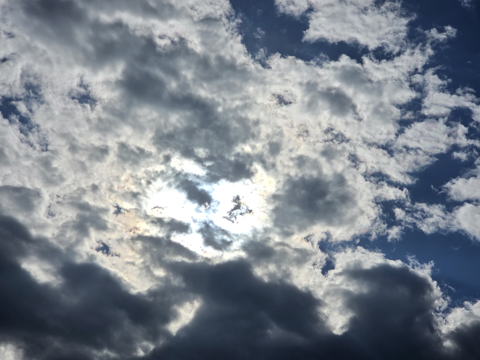 This media file is a photograph of the sky, featuring a dynamic interplay of dark and light clouds. The sun is partially obscured by thicker clouds, creating bright, hazy areas and casting shadows. The sky is a deep blue in some sections, contrasting with the white and gray of the cloud formations. The overall impression is of a dramatic and somewhat moody sky, possibly indicative of changing weather. There are no people, objects, text, or clear location cues to suggest a specific event or time of day beyond what can be inferred from the sky itself. The lighting suggests it could be daytime, with the sun making its presence known through the cloud cover.