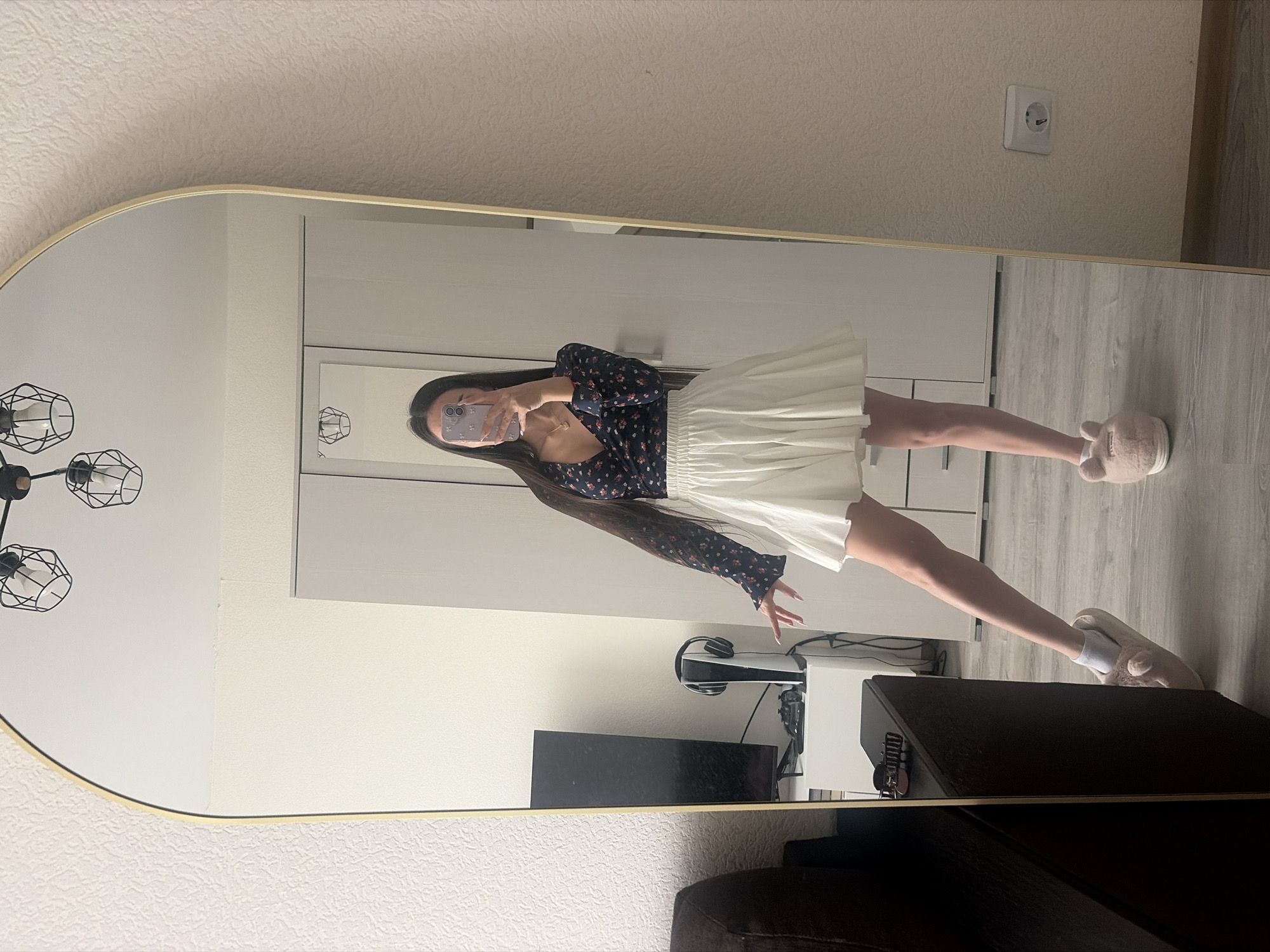 A young woman is captured in a full-length mirror selfie, standing upright in what appears to be a bedroom or living area. She is wearing a dark blue long-sleeved top adorned with a small floral print, paired with a white pleated mini-skirt. Her long dark hair cascades over her shoulders. She holds a light purple smartphone with a white case to take the picture, partially obscuring her face. On her feet, she wears white socks and light pink, fuzzy animal-ear slippers.
The setting is an indoor space with light-colored, possibly textured, walls and light wooden or laminate flooring. Behind the woman, a large white cabinet or wardrobe is visible. To her left, reflected in the mirror, are a black industrial-style chandelier with exposed bulbs and a section of a room containing a dark rectangular object and a white gaming console (likely a PlayStation 5) with headphones resting on top. A European-style electrical outlet is visible on the wall in the upper right of the reflection, consistent with the location context of Saint Petersburg, Russia. The mirror itself has a curved top and a gold or brass-colored frame. Part of a dark brown couch or chair is visible in the immediate foreground, reflected in the bottom of the mirror. The scene depicts a casual, private moment in a modern home environment. There is no visible text.