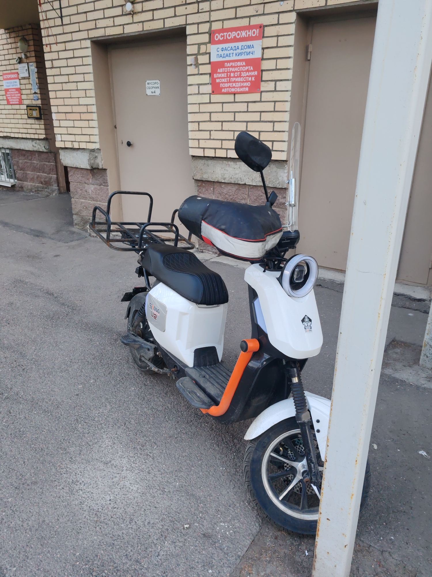 A white and black electric scooter is parked outdoors, angled toward the viewer, against a light brown brick building. The scooter features a large, black metal cargo rack above the rear wheel, a two-person black seat, and distinctive orange footrests. Its front has a circular LED headlight and a white fairing with a "STRONG" logo and "U2" branding. A white pole partially obstructs the front wheel and part of the scooter's right side.
The background consists of a brick wall with two doors. The door on the left is a darker beige, and an intercom system is mounted on the wall nearby. The door on the right is lighter brown and has a sign that reads "МУСОРО-СБОРНИК N4" (GARBAGE COLLECTION POINT N4). Above this door and to its left, a prominent red warning sign is affixed to the brick wall. This sign states "ОСТОРОЖНО!" (CAUTION!) and further warns in Russian: "С ФАСАДА ДОМА ПАДАЕТ КИРПИЧ!" (BRICK FALLING FROM THE FACADE OF THE HOUSE!) and "ПАРКОВКА АВТОТРАНСПОРТА БЛИЖЕ 7 М ОТ ЗДАНИЯ МОЖЕТ ПРИВЕСТИ К ПОВРЕЖДЕНИЮ АВТОМОБИЛЯ" (PARKING VEHICLES CLOSER THAN 7 M FROM THE BUILDING MAY LEAD TO DAMAGE TO THE CAR).
The ground is asphalt. The scene is brightly lit, indicating daytime and clear weather. No people are visible, suggesting the scooter is stationary and unoccupied. The setting is an urban environment, likely a side entrance or utility area of a building in Saint Petersburg, Russia.