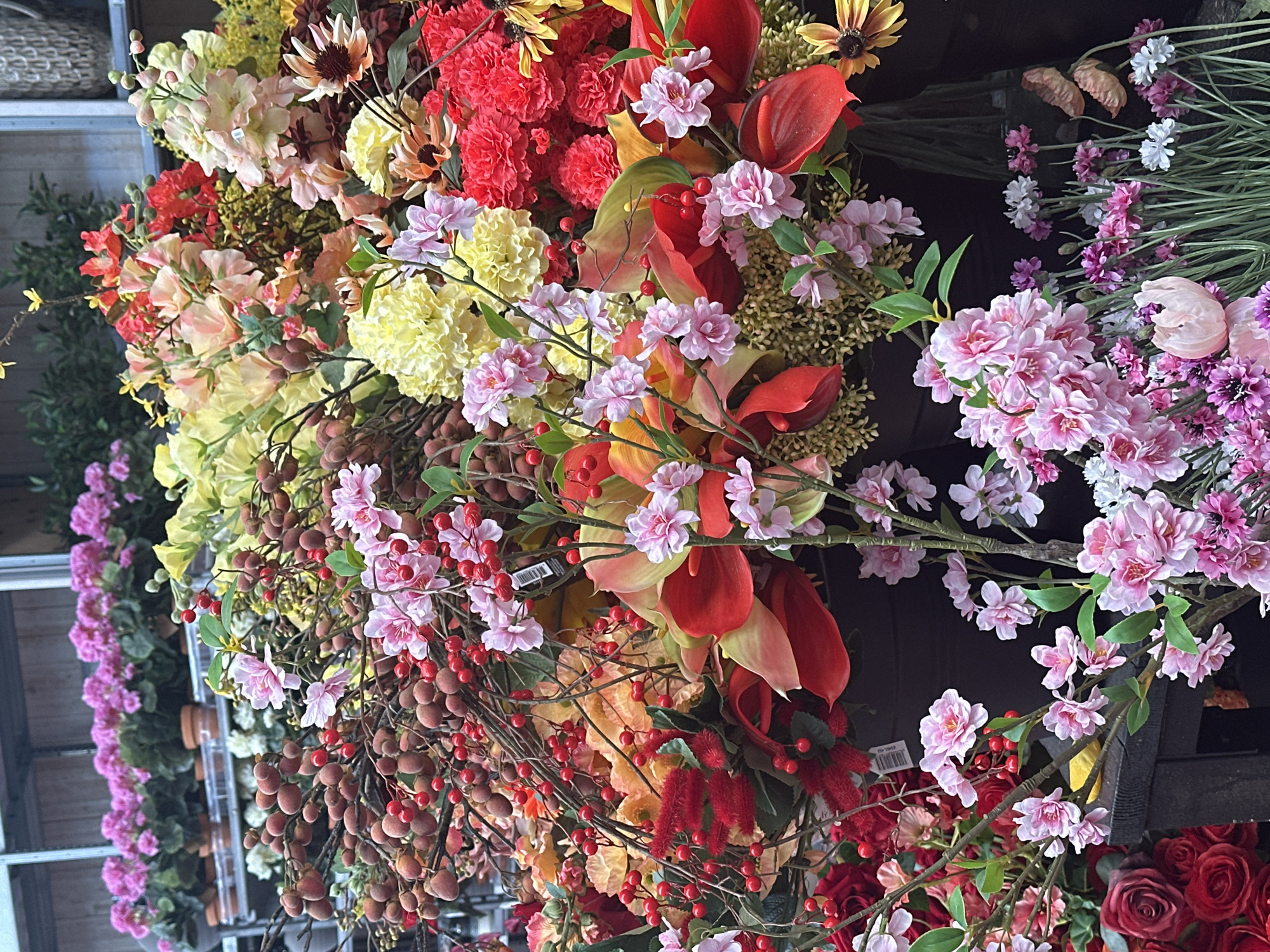 The image displays a close-up of a vibrant and abundant arrangement of artificial flowers, likely in a retail or decorative setting. The foreground is dominated by clusters of pink and red berries on thin brown stems, intertwined with various flowers in shades of pink, yellow, red, and orange. Notable floral elements include dense red carnation-like blooms, delicate pink blossoms, and larger, trumpet-shaped red and green leaves. Some branches with small, round, reddish-brown fruits are also visible. In the background, rows of potted pink flowers are seen on shelves, suggesting a garden center or florist shop. The overall impression is one of rich texture and color, characteristic of a floral display. There is no discernible indication of the specific location in Tuk, Netherlands, within the visual information provided.