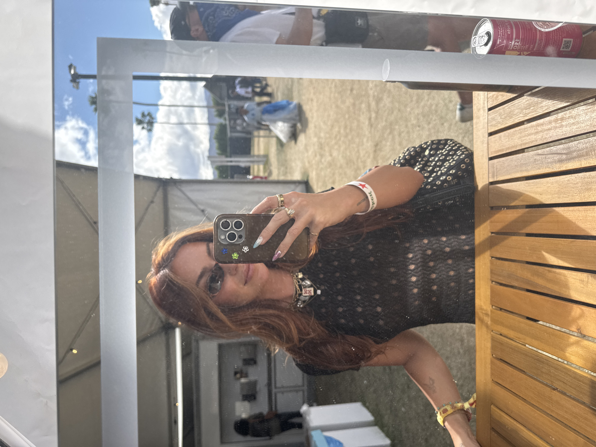 The image depicts a woman taking a selfie in a mirror. The mirror appears to be located in an outdoor setting, possibly at an event or festival. The reflection shows a glimpse of what looks like a tent, a grassy area, and other people in the background. The weather seems sunny, with a clear blue sky with clouds reflected in the mirror, along with palm trees. In the upper part of the mirror, a can of what looks like sparkling water or soda is placed on a ledge. The woman is wearing a black top, jewelry, sunglasses, and is holding a phone with multiple rings. Her hair is reddish-brown, and she has decorated nails. The image includes the reflection of what appears to be a wooden table or bench, and parts of what looks like a building.
Based on the presence of tents, palm trees, and other details in the mirror, it's likely that the location is in La Quinta, as it's known for hosting events like Coachella and Stagecoach music festivals.