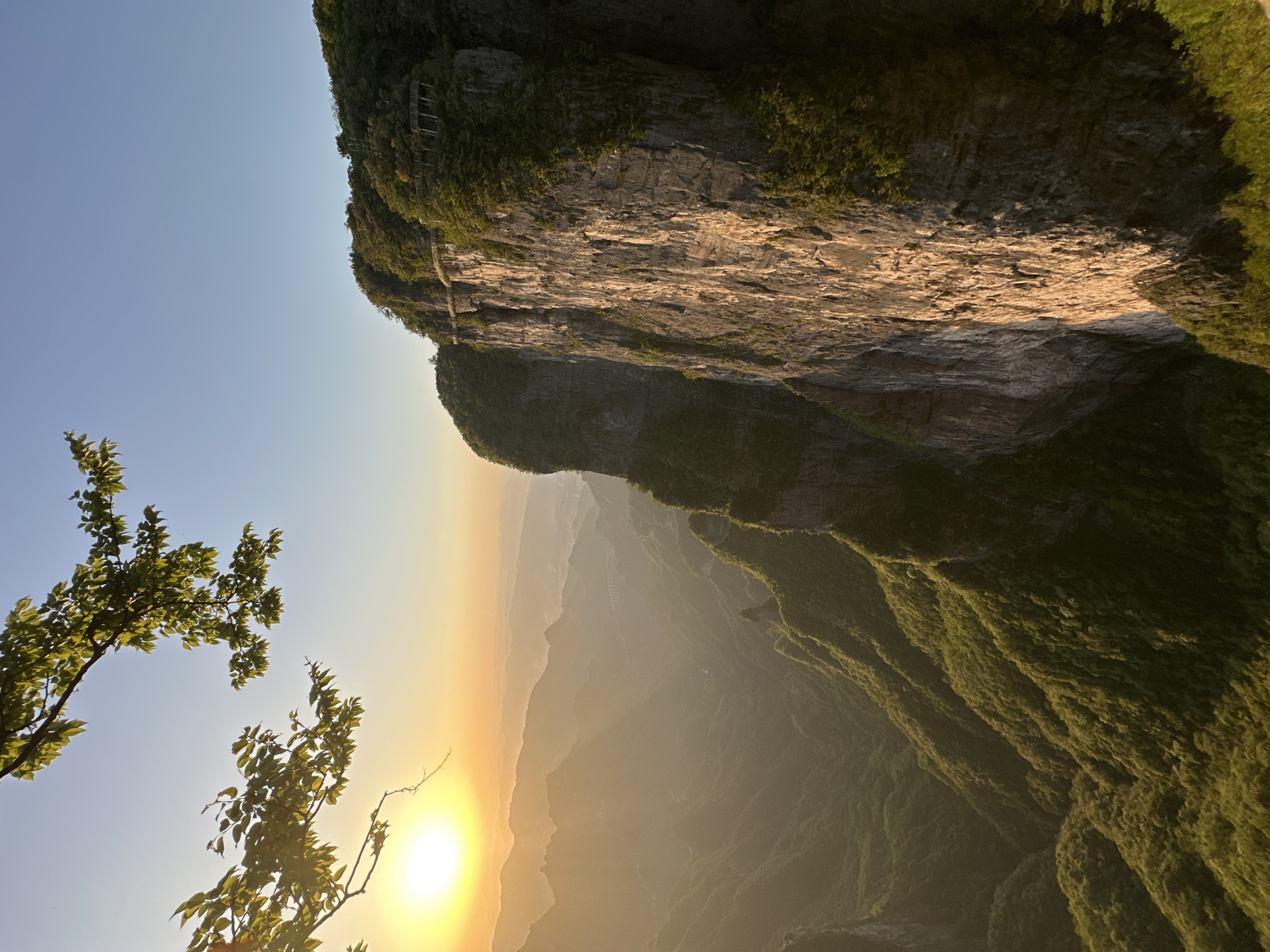 The image shows a landscape in Yongding District, China. The photograph is taken from a high angle, looking down on a steep, moss-covered cliff face. Part of a skywalk is visible near the top of the cliff. Below, layers of mountains are visible through a hazy, sunlit atmosphere. A tree is visible in the foreground on the left. The sun is positioned low in the sky, creating a warm, golden light.