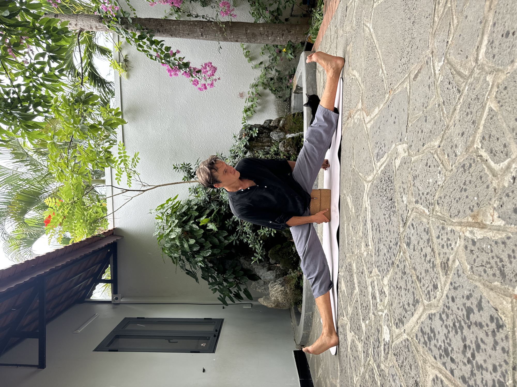 A man is performing a yoga pose outdoors in what appears to be a courtyard or patio. He sits on a white yoga mat on a stone-paved surface. He is wearing a black shirt and gray pants. The man has his legs extended wide apart in a straddle position. A wooden yoga block is placed under his seated area to assist the pose.
Behind the man is a white wall with various plants and greenery. A small fountain is also visible. A tree with pink flowers grows near the wall. The corner of a building with a roof and a window is visible to the left of the man.
The scene seems to be captured in Mũi Né, Vietnam, based on the architecture and vegetation.
