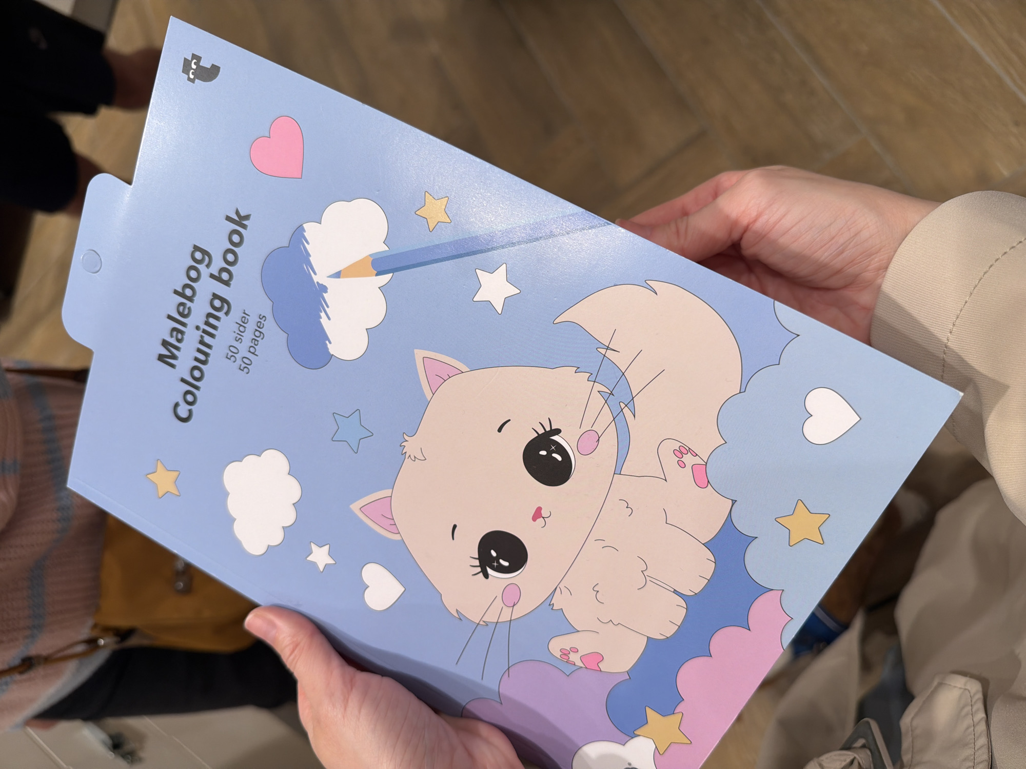 The image shows a light blue "Malebog Colouring book" with a cat illustration on its cover, held by two hands. The book, which claims to have "50 sider 50 pages", has a heart and star design on its cover, along with clouds and a pencil drawing. The background includes parts of people and what appears to be a beige jacket. The precise location in the city of Unknown, Unknown cannot be determined from the image.