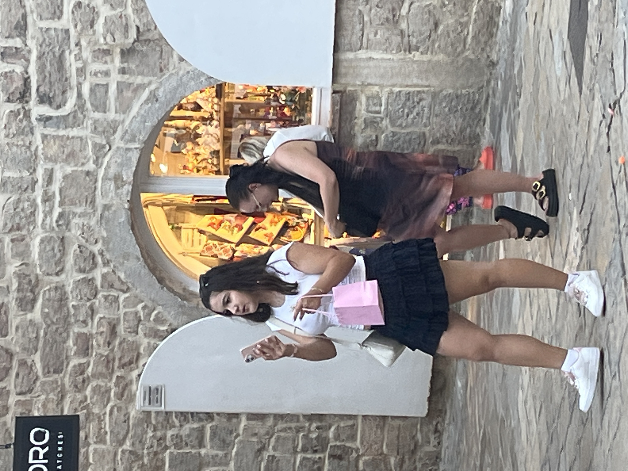 In Budva, Montenegro, two young women are seen walking in front of a store with a stone facade. The woman on the left, wearing a white t-shirt and a black ruffled skirt, holds a pink gift bag and a smartphone. She is wearing white sneakers. The woman on the right, dressed in a brown and black tie-dye dress, carries a black bag and wears black sandals. She is also wearing glasses. Behind them, a store window displays various items, possibly souvenirs, with colorful decorations. The ground is paved with cobblestones. A sign with black lettering that reads "ORO WATCHES" is visible on the upper left side of the image, attached to the stone wall.