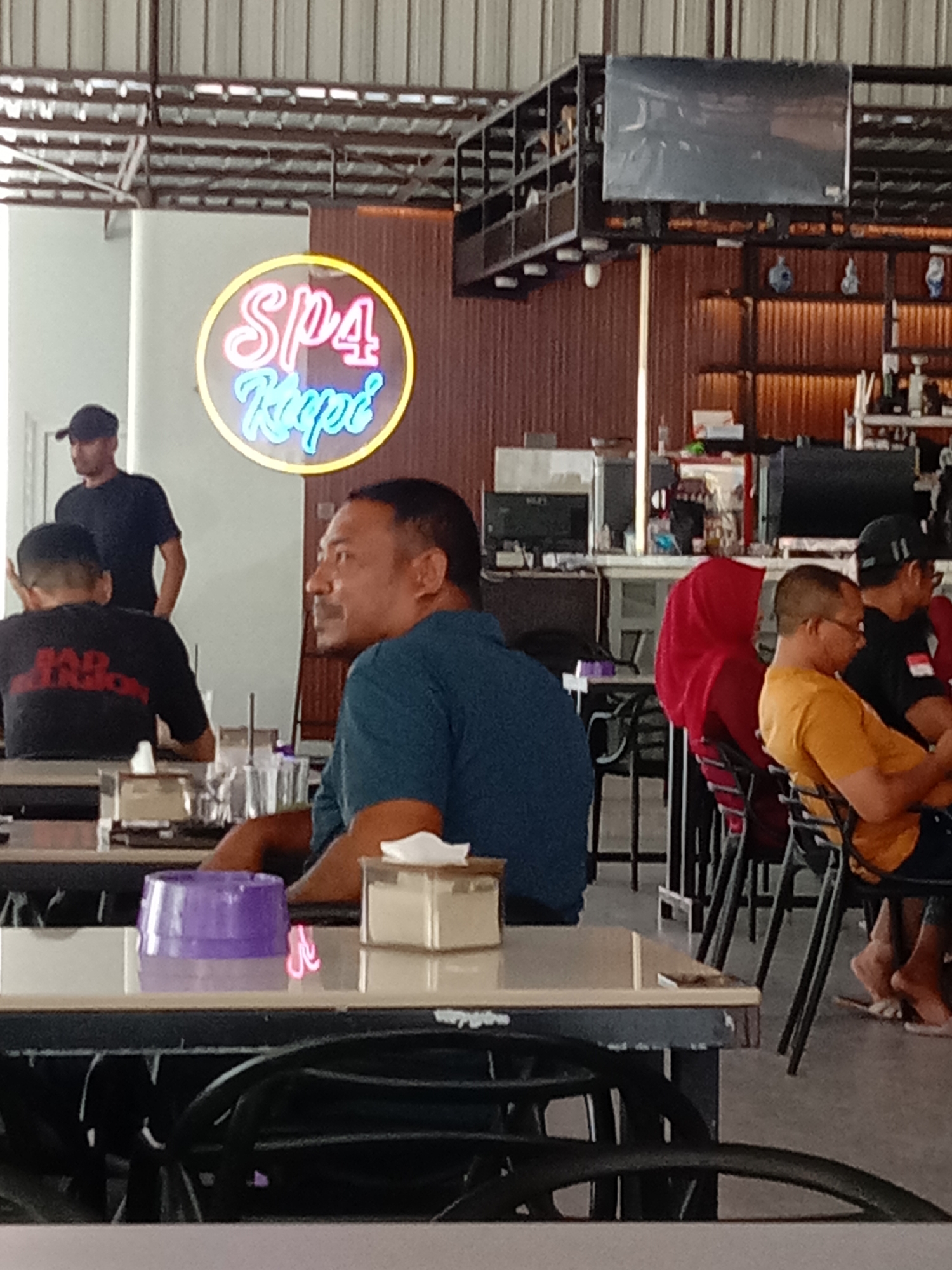 The image depicts the interior of a cafe or restaurant, possibly in Bireuen, Indonesia, as indicated by the neon sign that reads "Sp4 Kopi". In the foreground, a man with dark skin and short hair, wearing a blue t-shirt, is seated at a table, looking to his right. On the table in front of him are a purple container, a small white box, and a glass with some liquid. To his left, another man is seated with his back to the camera, wearing a black t-shirt with red lettering that appears to say "BAD TENSION". Further in the background, to the left, another man wearing a black t-shirt and a dark cap stands, looking towards the left.
The cafe's ambiance is suggested by the industrial-style ceiling with exposed metal beams and corrugated metal siding on the walls. Shelving units with various items, including what appear to be bottles and decorative objects, are visible in the background. A television screen is mounted on a shelf unit. Several other patrons are seated at tables in the background, including a person wearing a red headscarf and another wearing a yellow t-shirt. Chairs are arranged around the tables, and the floor is a light gray concrete. The lighting appears to be a mix of overhead lights and the neon glow of the sign.