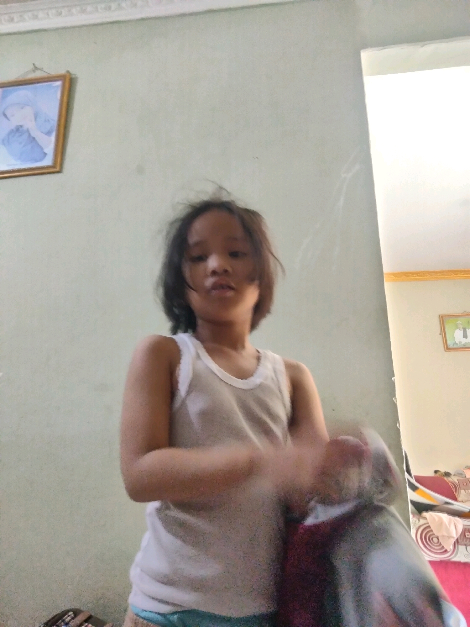 In Suka Sari, Indonesia, a young child stands in what appears to be a home environment. The child, with dark, messy hair, wears a thin white tank top. The background features a light green wall, with two framed pictures hanging on it. To the right, a doorway leads to a brightly lit area. The child's arms are blurred, suggesting movement, and they are holding an object that is also indistinct. A couch with patterned cushions is visible in the background on the right.