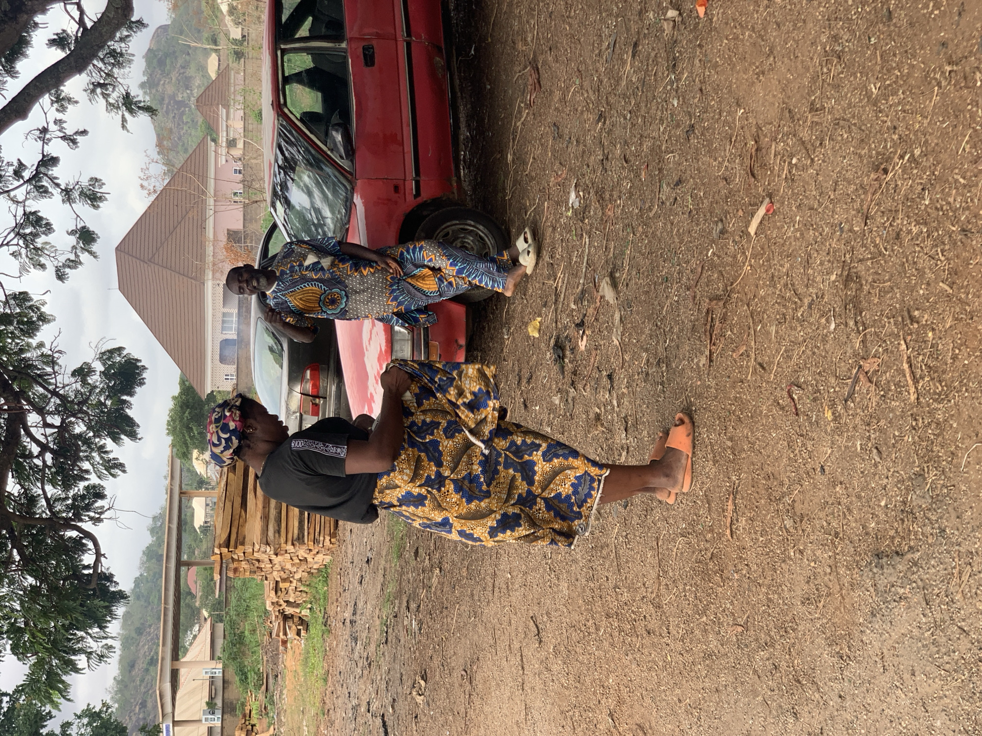 The image shows two people standing near a red car in Kabba, Nigeria. The ground is dirt and gravel. In the foreground, a woman wearing a black t-shirt and a blue and gold patterned skirt is standing. She is also wearing orange sandals and a patterned headscarf. Behind her, there is a stack of lumber. A man wearing a blue and yellow patterned shirt is leaning against the car, next to the woman. A house with a pyramidal roof is visible in the background, as are some trees and a distant hillside. The sky is overcast.