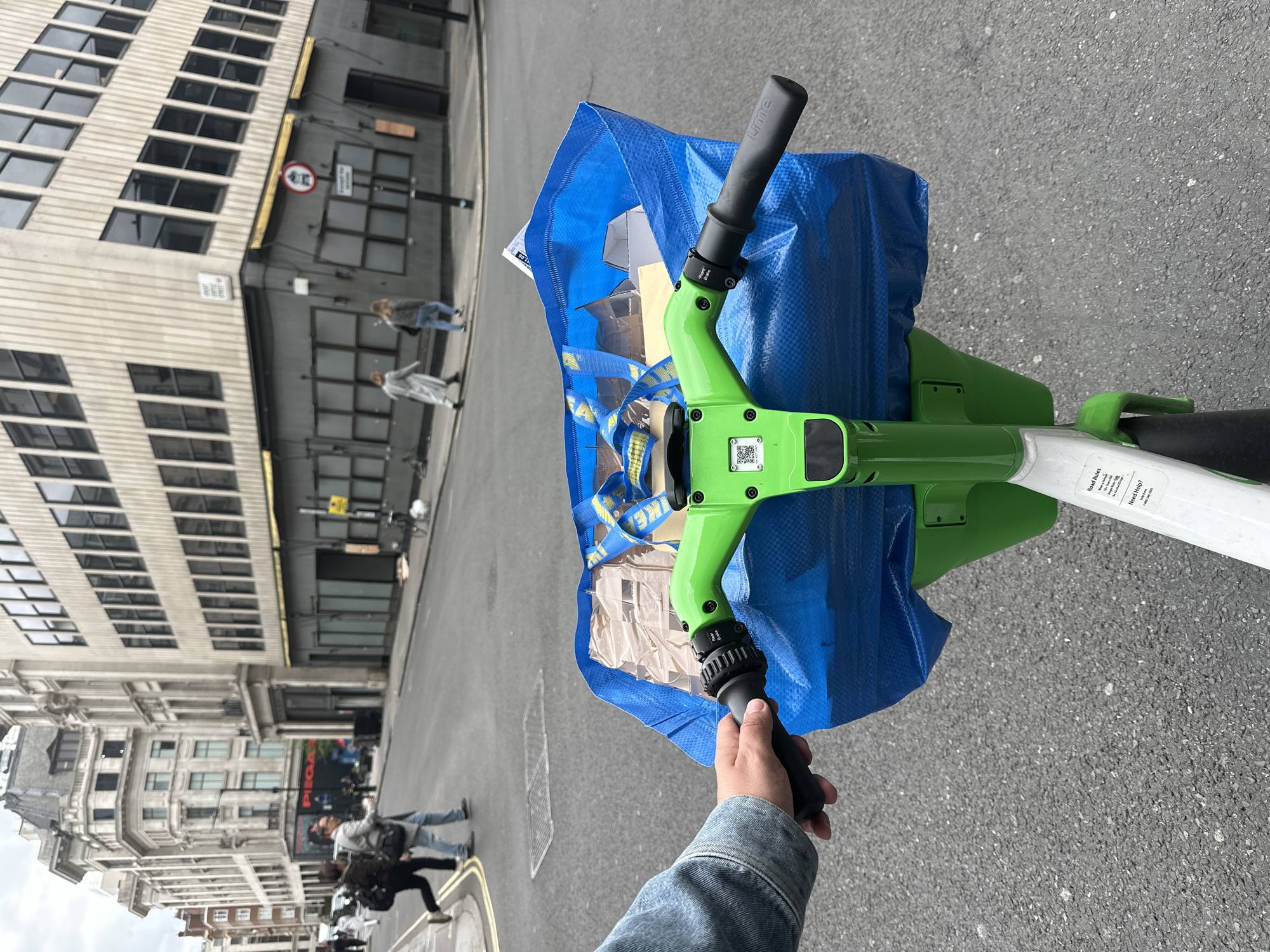 Here is a concise description of the image:
The image is taken from the first-person perspective of someone holding the handlebar of a Lime electric scooter. A blue IKEA bag filled with items is secured around the scooter's handlebar. The scooter is on a street in London. Pedestrians walk along the sidewalk, and buildings line both sides of the street. A street sign is visible in the background. The photo appears to have been taken during the daytime.