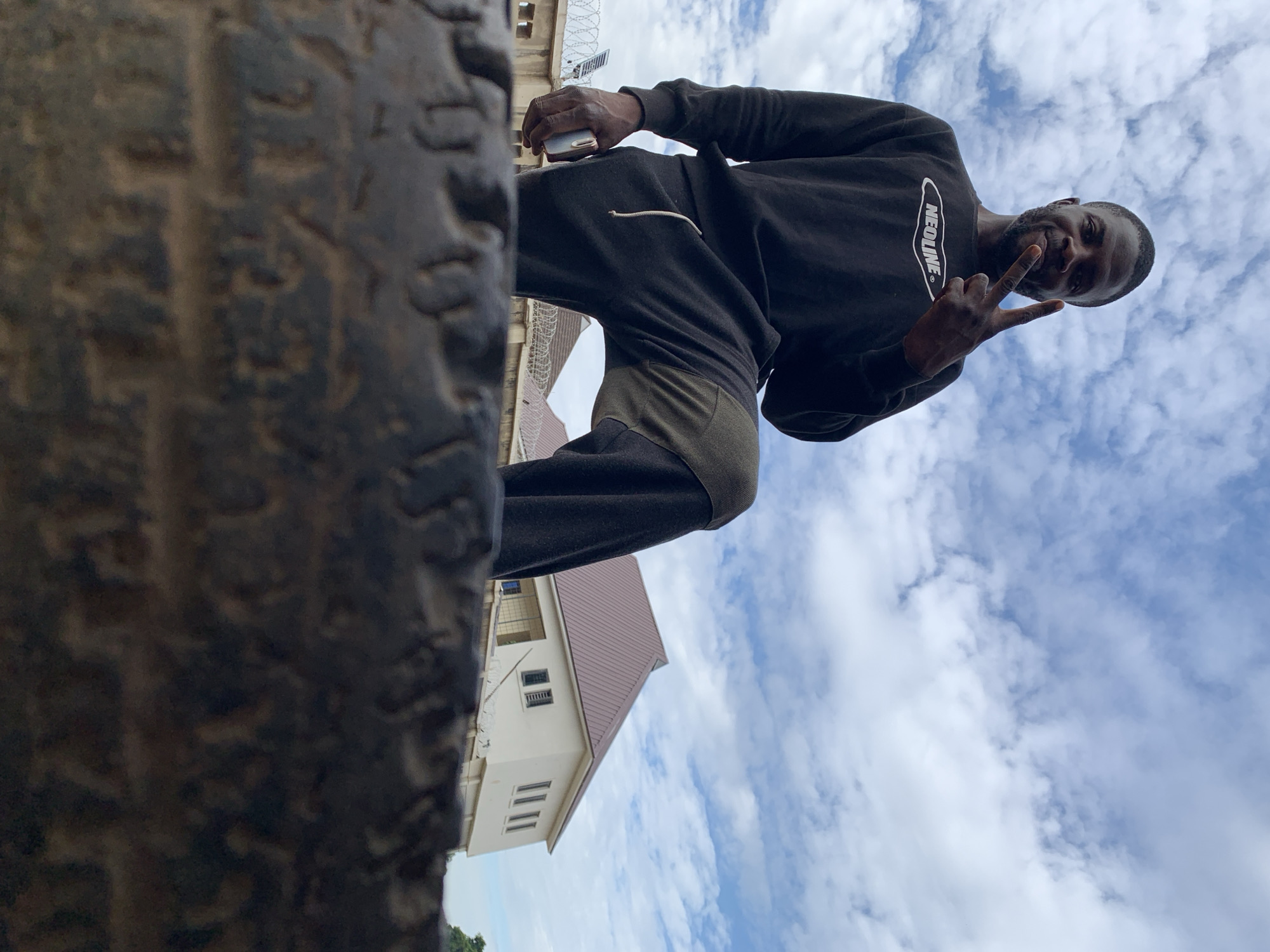 This image, captured in Rafin Par, Nigeria, depicts an adult male posing outdoors under a partially cloudy blue sky. The camera is positioned at a very low angle, looking upwards, with the immediate foreground occupied by a large, dark, textured object, appearing to be an old rubber tire, which obscures the ground.
The man, of dark complexion with short dark hair and a beard, is positioned centrally. He wears a black long-sleeved sweatshirt with a white "NEOLINE" logo across the chest, and dark grey sweatpants featuring a lighter drawstring and an olive-green fabric patch on the right thigh. He stands with his right knee raised, foot out of frame, and his left foot flat on the obscured surface. His left hand, holding a silver-colored smartphone, rests beside his left hip. His right hand is raised to shoulder height, with the index and middle fingers extended in a V-shape, forming a "peace" sign. He is looking directly at the camera with a slight smile.
In the mid-ground, behind the man, a cream-colored, two-story building with a reddish-brown gabled roof is visible. The building features multiple rectangular windows with dark frames. A fence topped with barbed wire or razor wire runs horizontally across the mid-ground, separating the man's immediate vicinity from the background structures. To the far left, a light-colored structure, possibly a satellite dish or antenna, is visible above the fence line. A glimpse of green foliage is present on the far right. The upper two-thirds of the image are dominated by the sky, which shows scattered white cumulus clouds against a light blue expanse.