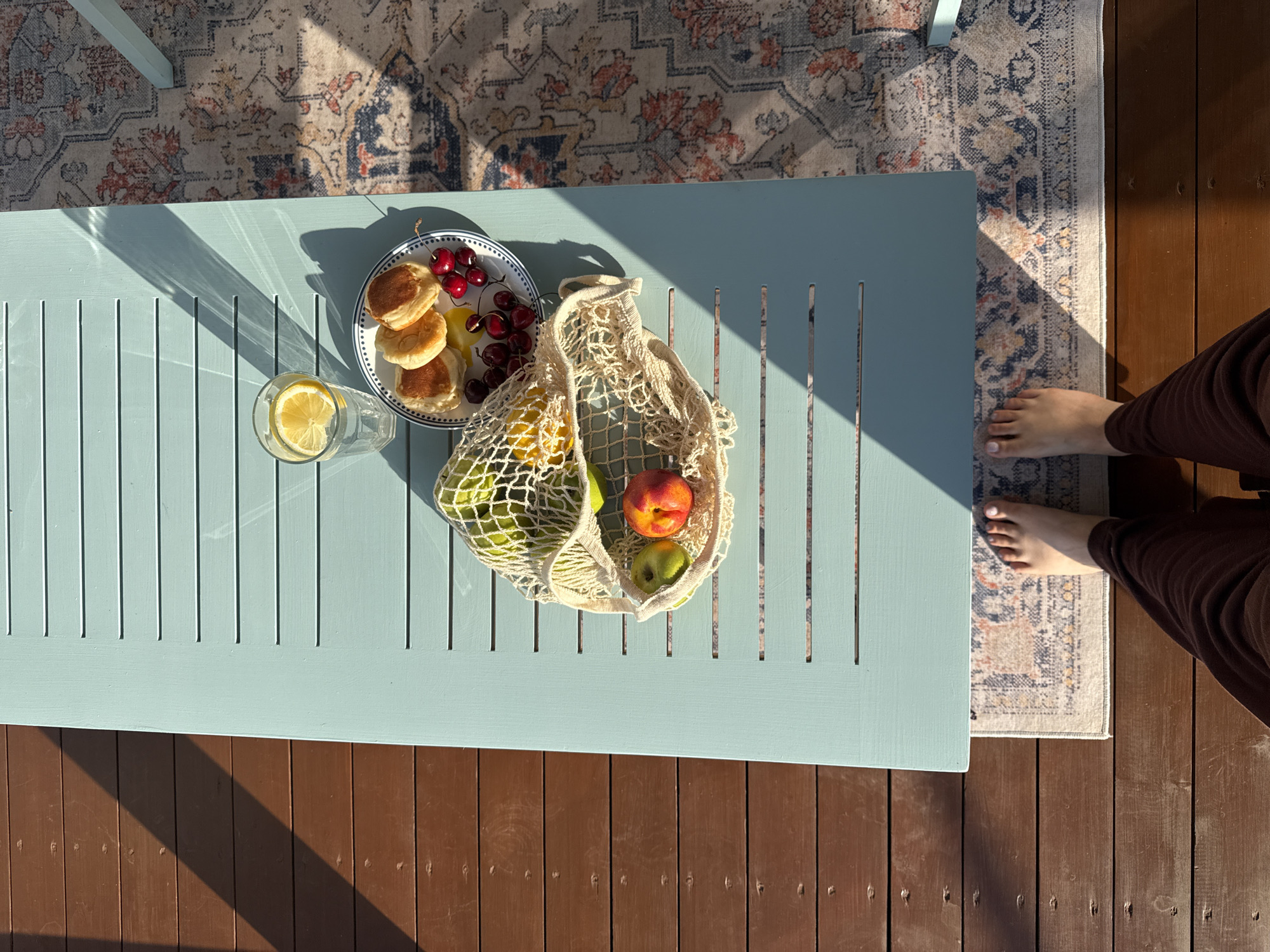 In this overhead shot of a light blue wooden table, a spread of breakfast items and fruit is bathed in warm sunlight, suggesting it is daytime. On the table, a plate holds several small, golden pancakes and a scattering of bright red cherries. Adjacent to the plate is a clear glass filled with water and a slice of lemon. A woven net bag rests near the plate, filled with green apples, a nectarine, and possibly other fruits. The table is situated outdoors, as indicated by the shadow of a rug and the presence of wooden decking beneath it. A patterned rug, partially visible, lies on the decking, and a person's bare feet and brown trousers are visible on the right edge of the frame, suggesting they are sitting at the table. The overall mood is relaxed and pleasant, with the natural light creating a serene atmosphere. The image appears to have been taken in Starokorsunskaya, Russia.