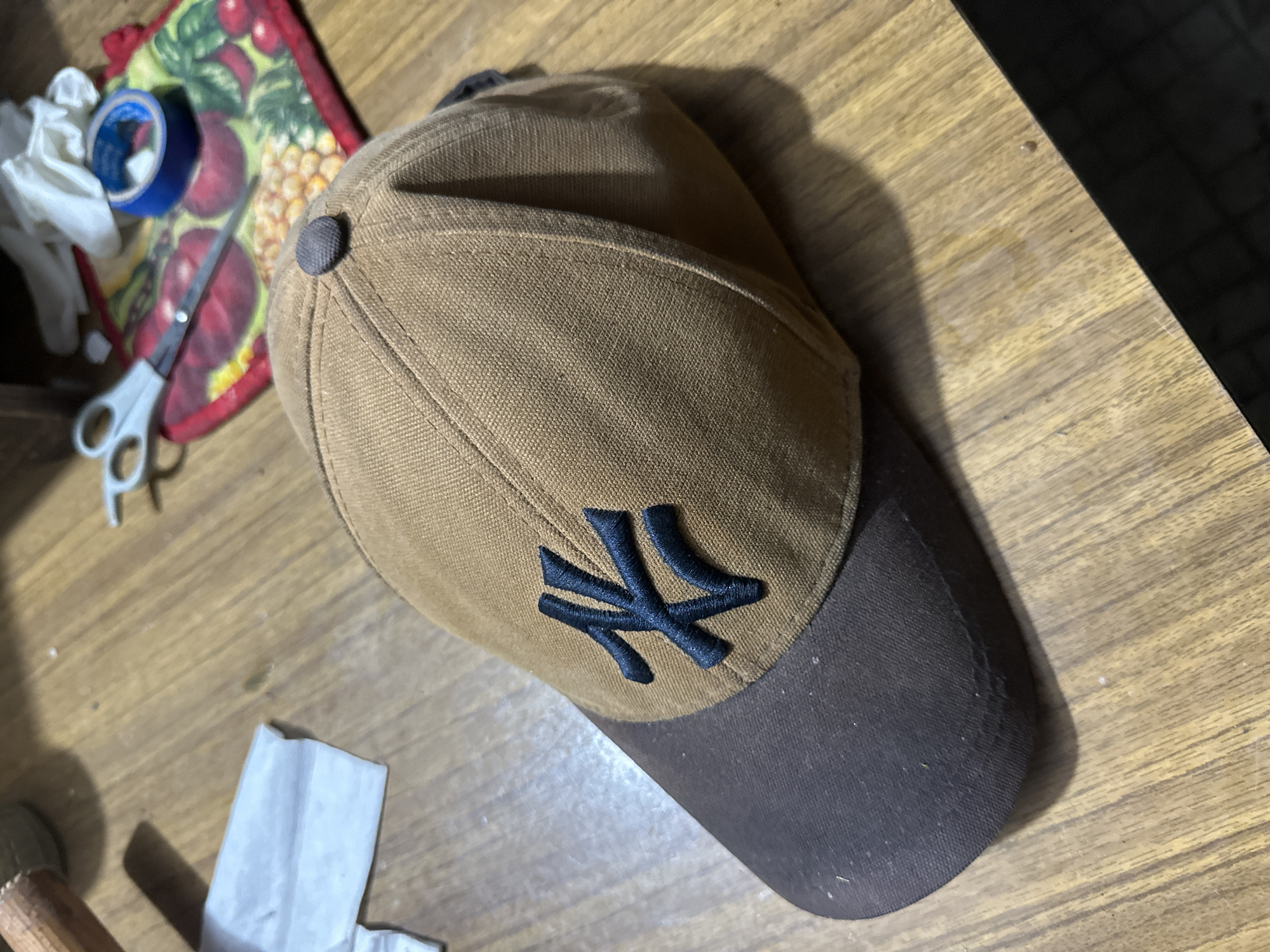 A close-up, eye-level, medium shot captures a tan baseball cap with a dark brim, emblazoned with a black "NK" logo, resting on a wooden table. The cap is positioned slightly off-center, with its front facing towards the viewer. The brim is dark brown, and the main body of the cap is a lighter tan color. The "NK" logo is a stylized black embroidery. A dark gray button sits atop the crown of the cap. The wooden table has a visible grain pattern. In the background, out of focus, are a white folded cloth, a pair of scissors, a roll of blue tape, and a colorful mat with fruit designs. The scene is well-lit, casting shadows from the cap onto the table. This image is not located in Pîrlița, Moldova.