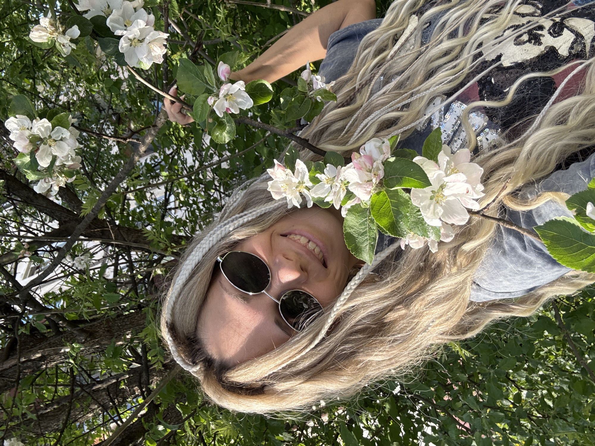 A woman with blond hair, accented with braids, wearing round sunglasses, is positioned amidst the branches of a flowering tree. She is smiling. White blossoms with green leaves surround her face and extend into the background. The background includes additional foliage and tree branches. Her clothing is visible, consisting of a gray top and a black t-shirt with sequins. Location: possibly Rusanovo, Russia.