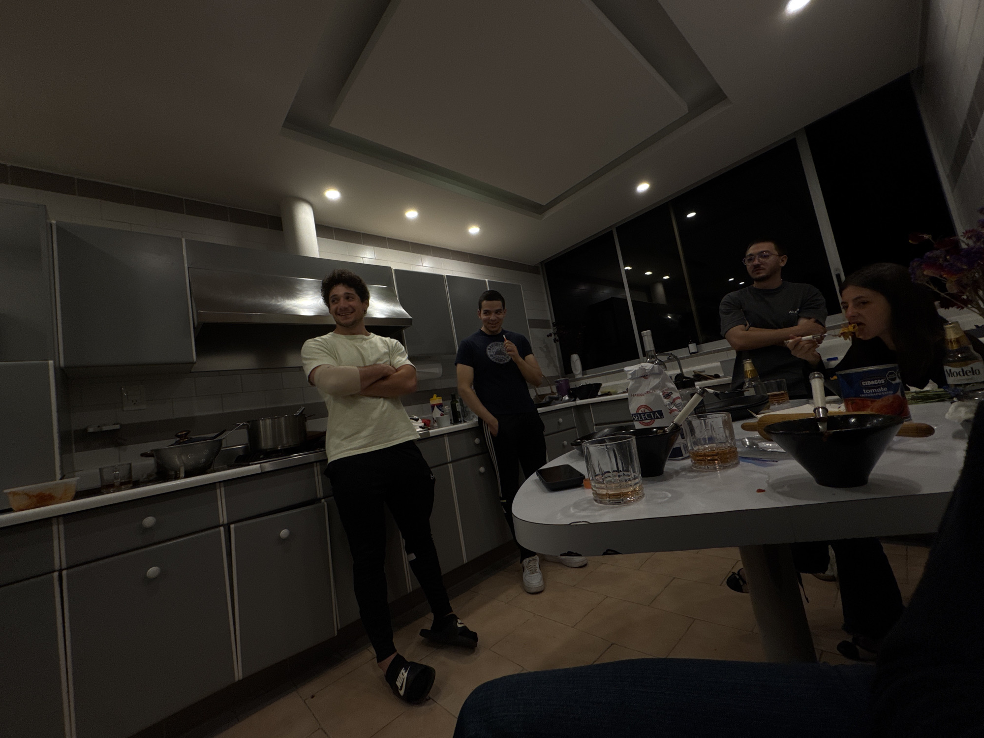 The image depicts a casual social gathering of four people in a modern kitchen and dining area during the evening or night, indicated by the dark windows overlooking what appears to be city lights. The setting features a contemporary kitchen with sleek dark gray cabinets, a stainless steel range hood, and light countertops, adjacent to a dining table.
On the left, a man with curly dark hair, wearing a light t-shirt and dark pants, stands with his arms crossed and a smile on his face. He is wearing a "Nike" slide sandal. Behind him, another man in a dark t-shirt stands, smiling while holding an object to his mouth. Further right, a man with glasses and a dark t-shirt stands with his arms crossed, observing. At the dining table, a woman with long dark hair is actively eating from a bowl with a fork. The leg and knee of a fifth person are visible in the lower right foreground, suggesting another individual seated at the table.
The table is adorned with several clear glasses containing an amber liquid, a black bowl of food, a white bag of "HARINA SELECTA" (flour), a red can labeled "CIDAGOS Tomate Triturado" (crushed tomatoes), a "Modelo" beer bottle, a rolling pin, and a vase with artificial flowers. Pots are visible on the kitchen stovetop. The overall mood appears relaxed and social, characteristic of friends or family enjoying food and drinks together in a convivial atmosphere. The visible product branding, such as "Modelo" and "Harina Selecta," aligns with the given Mexico City location context.