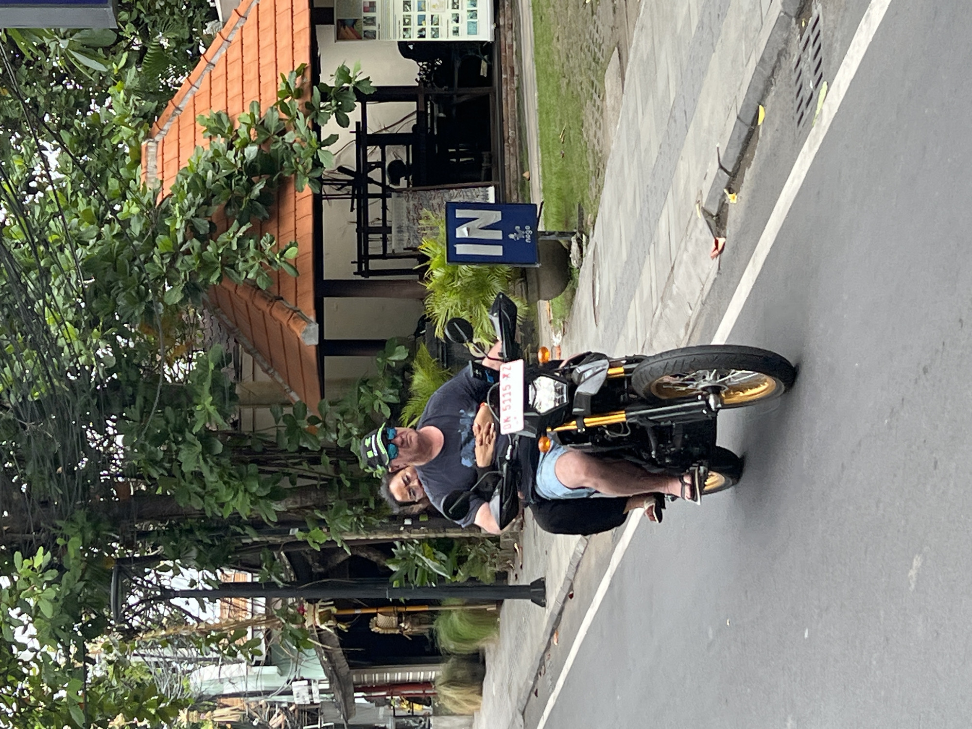 In Denpasar, Indonesia, a man and a woman ride a motorcycle on a paved street. The man, wearing a dark t-shirt, blue shorts, a green baseball cap, and sunglasses, drives the motorcycle. The woman, wearing a dark outfit, sits behind him, holding his waist. The motorcycle is black with gold accents. In the background, there are buildings with terracotta roofs, lush green trees, a lamppost, and a sign that says "IN" with a blue logo below it. The street has a white line marking the lane.