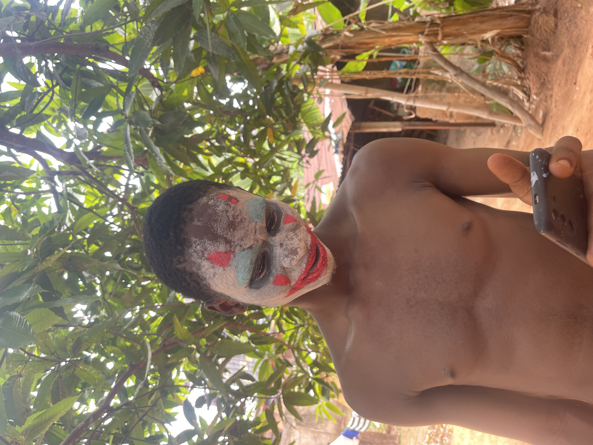 A shirtless, dark-skinned person with clown-like face paint is holding a phone in their right hand. The face paint consists of white base, red around the mouth, and blue triangles above and below the eyes. The person is standing outdoors beneath a tree with green foliage, with wooden structures and a red dirt ground visible in the background. The location is likely in Onitsha, Nigeria, but without further context, this is just an assumption.