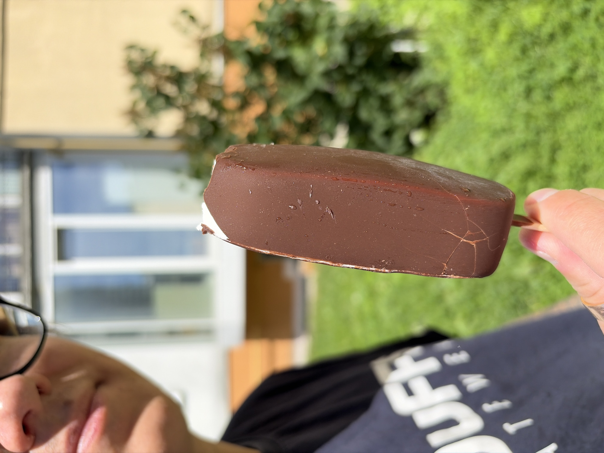 A close-up, outdoor, eye-level, medium shot shows a person holding a chocolate-covered ice cream bar in their right hand. The person is wearing glasses and a dark t-shirt with white lettering. The ice cream bar is partially melted, with water droplets on its surface. The background is blurred, showing a grassy area and a building. The lighting suggests it is a sunny day. The person's left shoulder and chest are visible, along with their mouth and nose. The overall scene depicts a person enjoying an ice cream on a warm day.