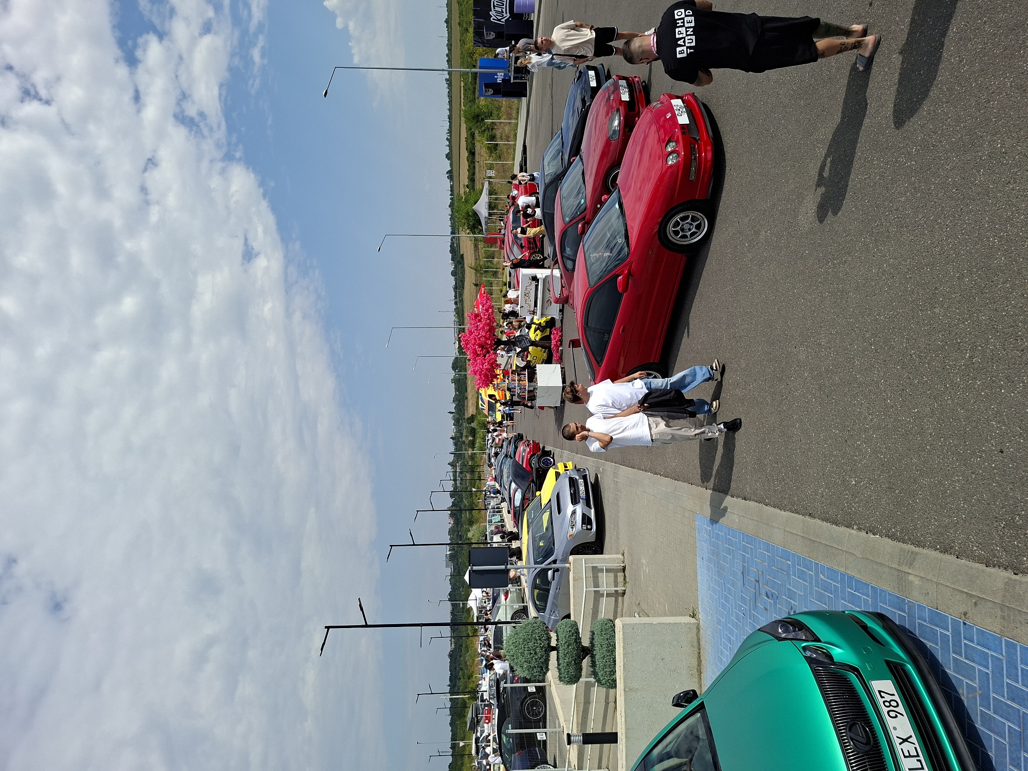 The image depicts an outdoor car meet event on a sunny day. In the foreground, a vibrant green Lexus GS is parked on a blue tiled surface, its license plate reading "LEX 987". To the right, two young men are walking away from the camera. The scene opens up to reveal a parking lot filled with various cars, many of which appear to be modified or sports cars. Several people are milling about, interacting and observing the vehicles. In the background, there are tents, some decorated with pink artificial trees, suggesting vendor stalls or event activities. Streetlights are visible throughout the parking area. The overall environment is flat with some vegetation and distant buildings visible on the horizon under a partly cloudy sky. Based on the provided information, this event is located in the city of Stăuceni, Moldova.