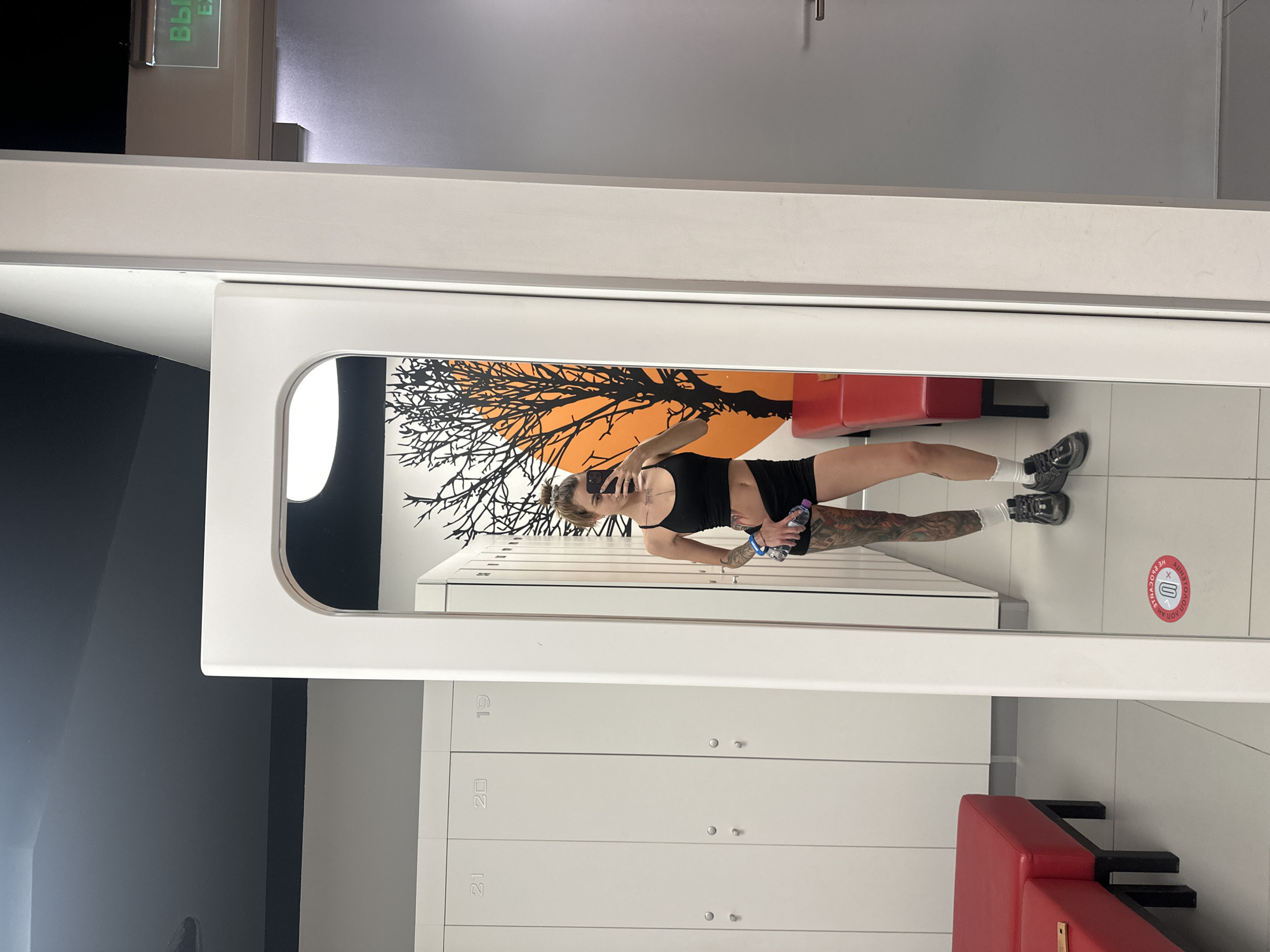 In a locker room, a person stands in front of a full-length mirror, taking a selfie with their phone. The person is wearing a black sports bra and black shorts, with white socks and athletic shoes. They have extensive tattoos on their left leg and arm. In their right hand, they hold a water bottle.
The locker room features rows of white lockers with numbers visible, such as 19, 20, and 21. Red benches are placed in front of some of the lockers. The wall behind the person in the mirror is decorated with a large mural of a tree silhouette against an orange sun.
To the right of the mirror, a gray door with a silver handle is visible. Above this door, a green illuminated sign reads "ВЫХОД," which translates to "EXIT."
The floor is tiled with light-colored squares. A circular red and white sticker with an arrow and text is on the floor in front of the mirror. The ceiling is dark gray. The scene suggests a fitness facility or gym. The presence of the "EXIT" sign in Russian indicates this location is in Russia, and the specific context of a gym locker room in Moscow, Russia, is plausible given the visual cues.