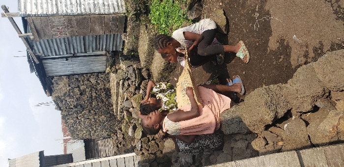 The image captures four young individuals, likely girls, engaged in an outdoor activity in the city of Buzove, Democratic Republic of the Congo. The scene unfolds on a dirt ground surface featuring exposed, dark, porous rock formations and constructed steps leading upwards into the background. Surrounding structures include buildings with corrugated metal walls and roofs, and a prominent wall built from dark, irregular stones. One metal structure on the far right has faded, illegible text on its surface. In the immediate foreground, two girls are seated on large dark rocks, jointly holding a light-colored, rectangular board or tablet with visible markings on its surface, suggesting writing, drawing, or a game. The girl on the left wears a peach-colored sleeveless dress and blue sandals, while the girl on the right wears a white short-sleeved top, dark leggings, and patterned light-colored sandals. A third girl, wearing a green and white patterned dress, is seated behind the left-most girl, looking upwards and to the right, with one hand touching her head. A fourth, younger child is partially visible kneeling or sitting to the right, also observing the board. The environment is illuminated by bright daylight, casting defined shadows.
