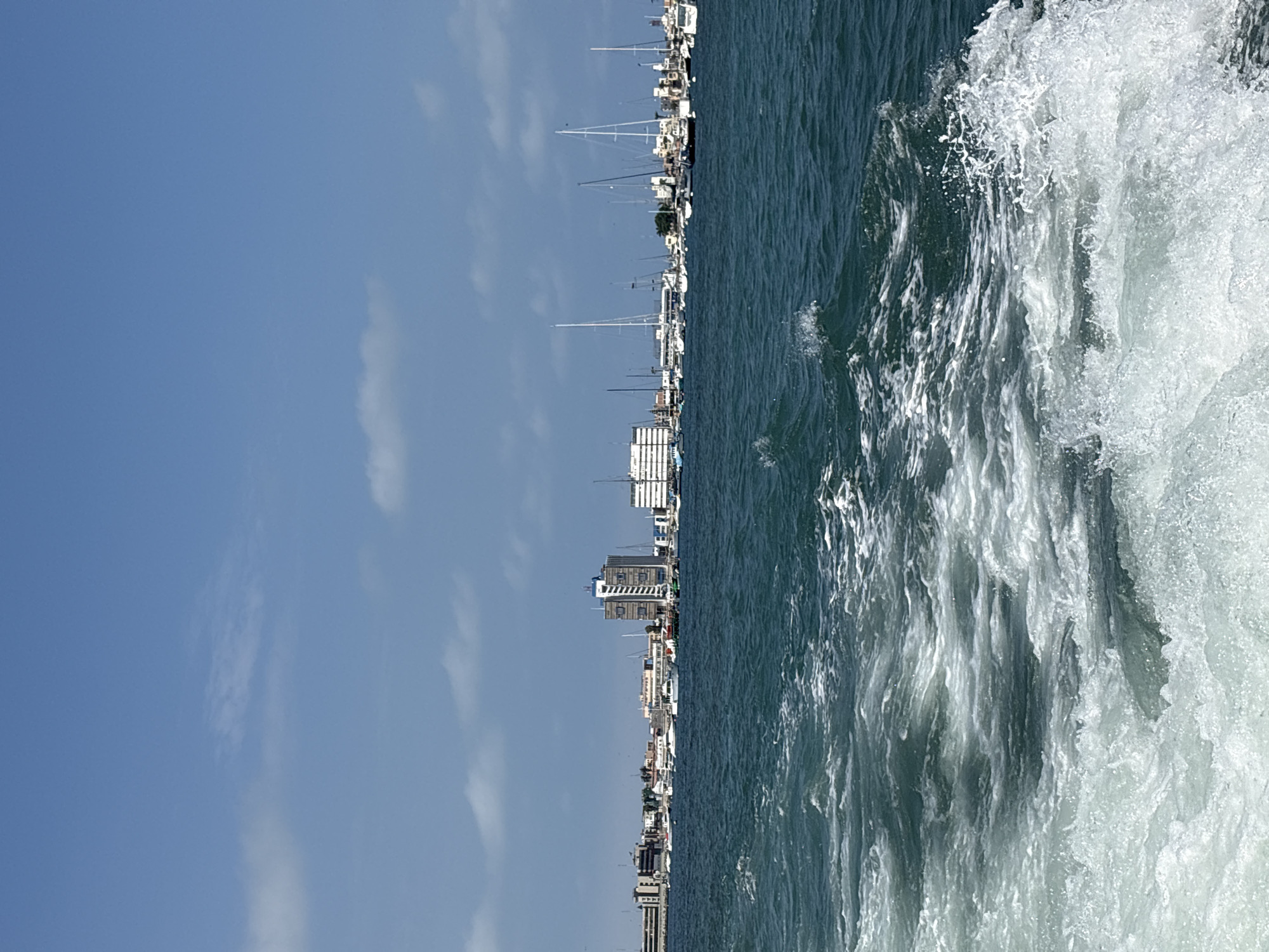 A body of water with a wake from a boat in the foreground, leading to a distant cityscape under a blue sky with scattered clouds. The cityscape features a marina with numerous sailboats and yachts, as well as several buildings, including a tall, modern high-rise. The water is a deep blue-green and appears choppy. The scene depicts a coastal urban environment, likely the port area of Callao, Peru, given the prominent maritime activity and the style of architecture. There are no people visible in the image.