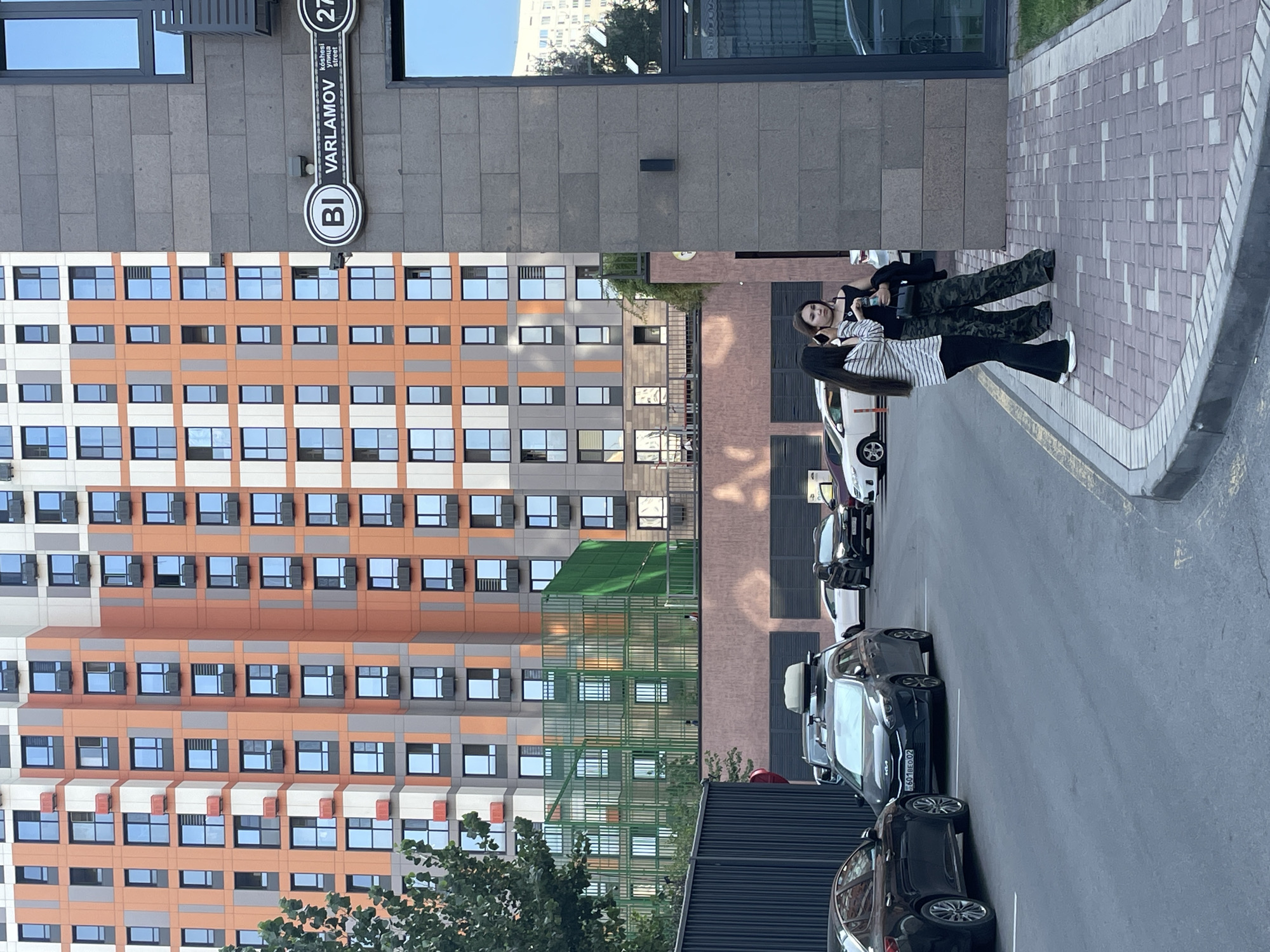 In Almaty, Kazakhstan, a residential building with orange and gray facade is visible. In front of the building is a parking lot with several cars. Two women are standing on the sidewalk in front of a building with a dark gray brick facade. One woman is wearing a black off-the-shoulder top and camouflage pants, holding a water bottle. The other woman is wearing a striped shirt and black pants. A street sign indicates "Varlamov street".
