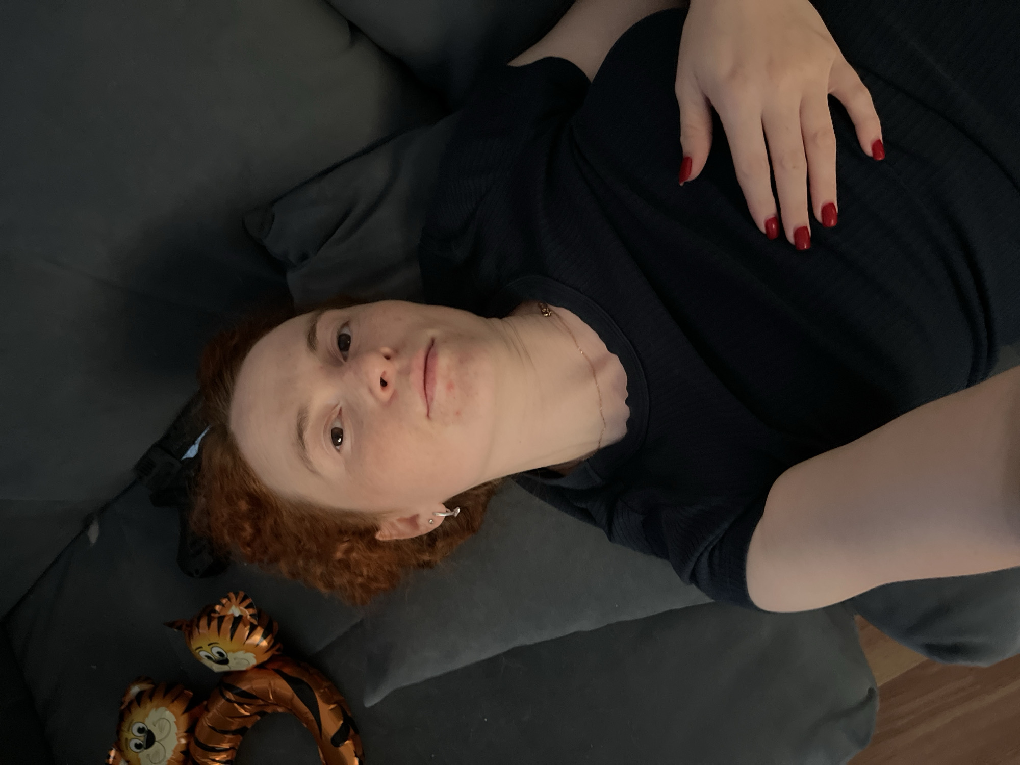 A person is lying on a gray couch. The person has red hair and is wearing a black shirt. Their hand is resting on their stomach, and their fingernails are painted red. A tiger balloon is on the couch next to the person's head. The location is identified as Chaikovsky, Russia.