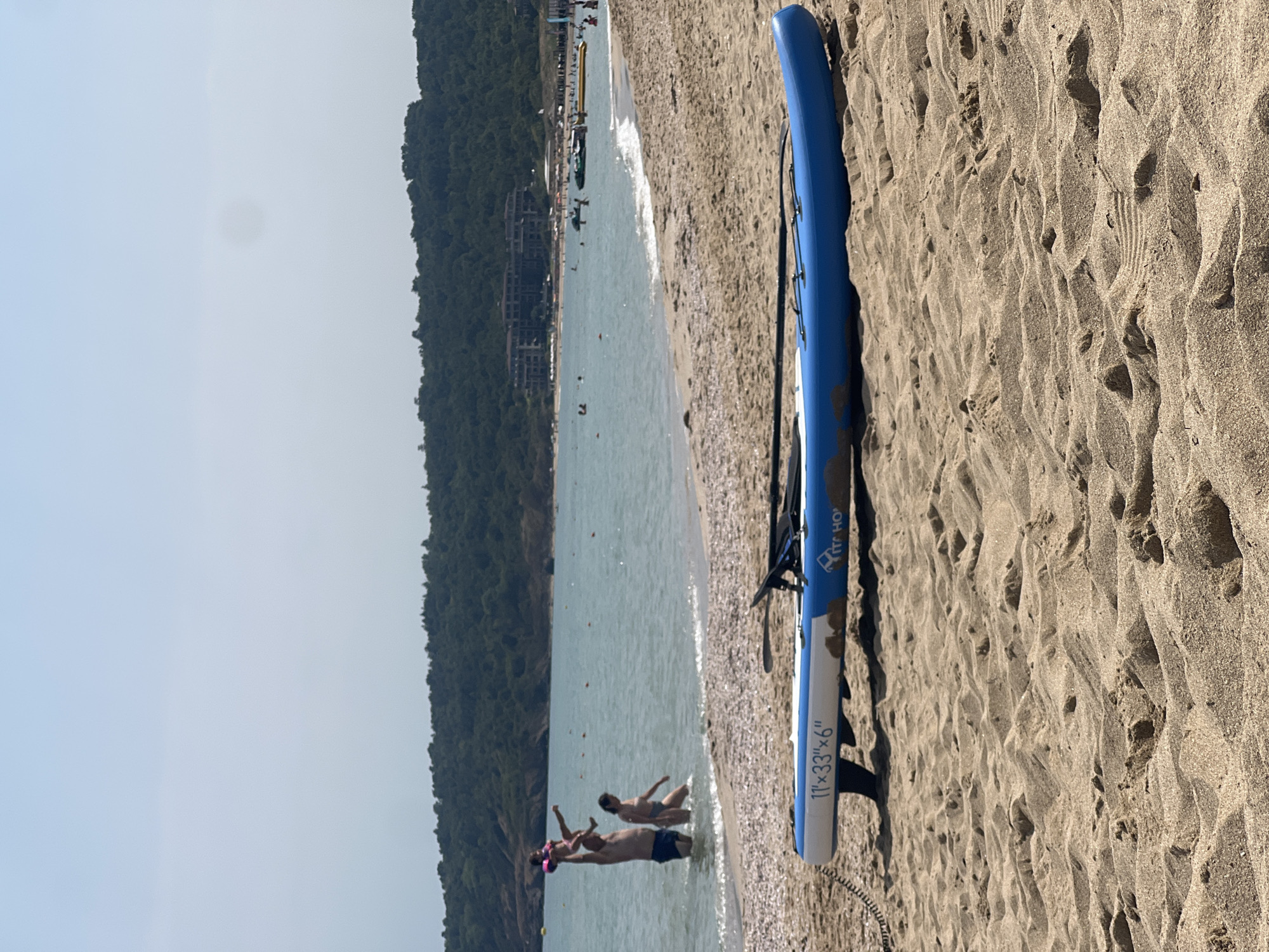 In Sozopol, Bulgaria, a blue and white paddleboard with the inscription "11'x33"x6" KITE HO" lies on a sandy beach, positioned diagonally in the foreground. The paddleboard has a black fin and a black paddle resting on top. Beyond the beach, the turquoise water of the Black Sea is visible, with several people wading and swimming. Further out, a family is engaged in play; a man is lifting a young child, who is wearing a pink life jacket, above his head while a woman stands nearby in the shallow water. In the background, a lush green, tree-covered hill rises, with buildings visible among the trees. The sky is clear and light blue with a slight haze.