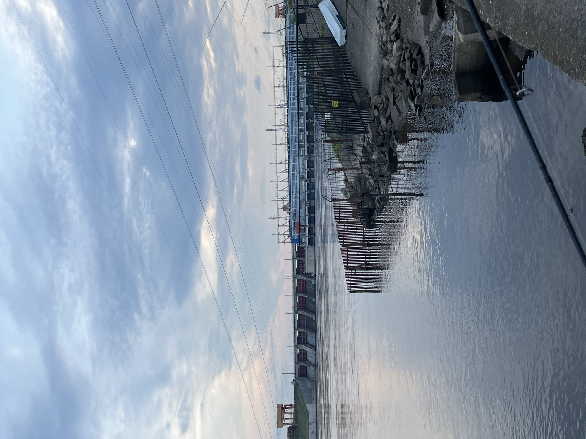 An expansive outdoor scene depicts a large industrial structure spanning a wide body of water under a partially cloudy sky. The main subject is a lengthy hydroelectric dam or power plant, characterized by numerous gates or sections, extending horizontally across the frame. Tall transmission towers and power lines rise from the structure, cutting across the sky.
In the foreground, on the right side, a concrete bank covered with loose rocks leads down to the water. A black fishing rod with a visible reel is cast into the water from this bank. A small, white, inverted boat or container rests on the concrete. A black metal fence runs along the bank, parallel to the dam, and a yellow sign with indistinct black text is affixed to it. The water below the dam shows ripples and reflects the overcast sky, suggesting calm conditions. The sky, which occupies the upper portion of the image, is filled with light, scattered clouds.
The lighting suggests either early morning or late afternoon, with soft, diffused light, indicating twilight hours. The location cues, particularly the scale and type of industrial facility, are consistent with a hydroelectric power plant, likely the Votkinsk Hydroelectric Power Plant near Chaikovsky, Russia. No people are visible.