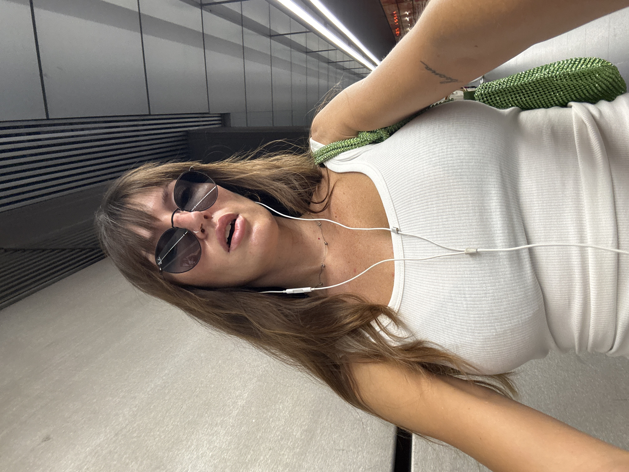 A woman with long brown hair, bangs, and sunglasses is looking into the camera in what appears to be a subway station. She is wearing a white ribbed tank top and has white earbuds in. A green sparkly purse is slung over her right shoulder. She has a tattoo on her right arm and a small necklace with red hearts. The background is a textured wall and a ceiling with horizontal lines.