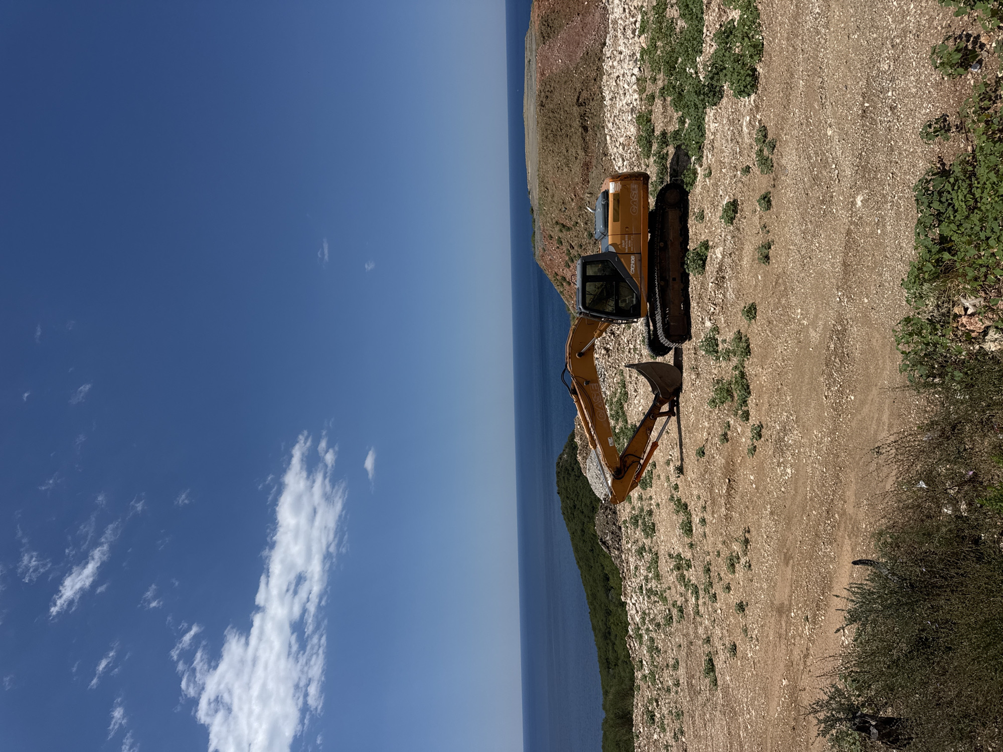 An excavator, identified as a CASE CX210B, sits idle on a rocky, dusty construction site overlooking the Adriatic Sea. The excavator is orange and black, with its boom extended and bucket lowered, appearing to be filled with gravel. The surrounding terrain is sparsely vegetated with low-lying green plants and scattered rocks. In the background, a lush green hillside slopes down to the deep blue sea, which stretches to the horizon under a bright blue sky dotted with scattered white clouds. The scene is likely located in the vicinity of Drobnići, Montenegro, a region known for its coastal beauty and ongoing development. There are no people visible in the image.