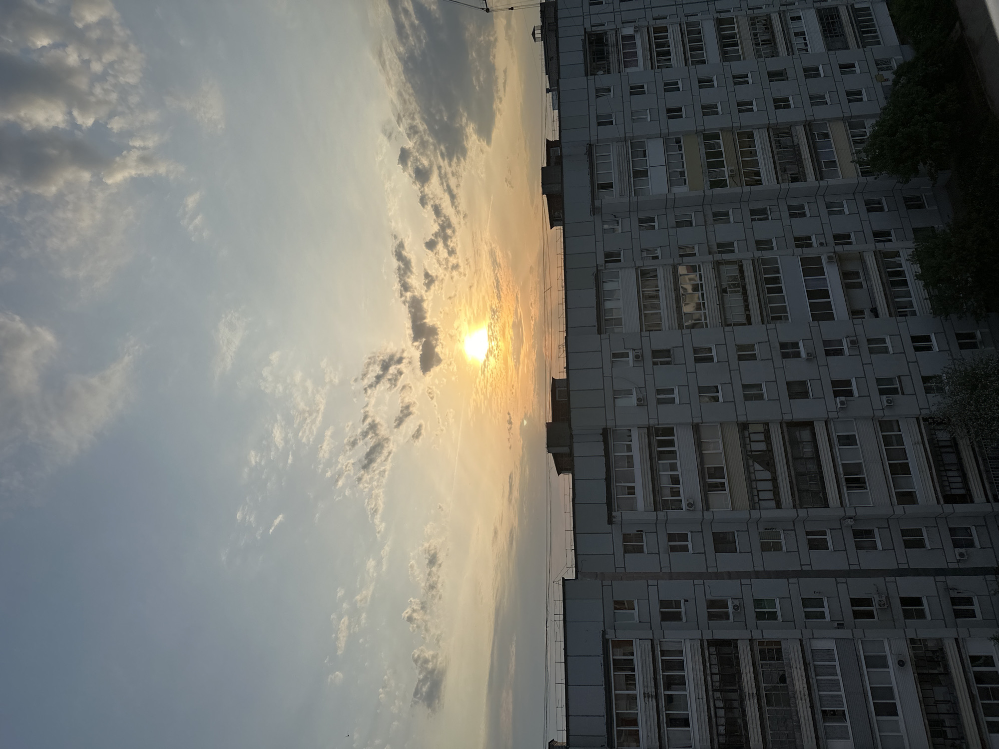 The image shows a building adjacent to a sky filled with clouds and the setting sun. The building is a multi-story, likely residential, structure with numerous windows and air conditioning units. The sky is filled with clouds, with the sun positioned in the center, partially obscured by the clouds, creating a hazy, bright light. The location is assumed to be in an urban area, possibly Chaikovsky, Russia.