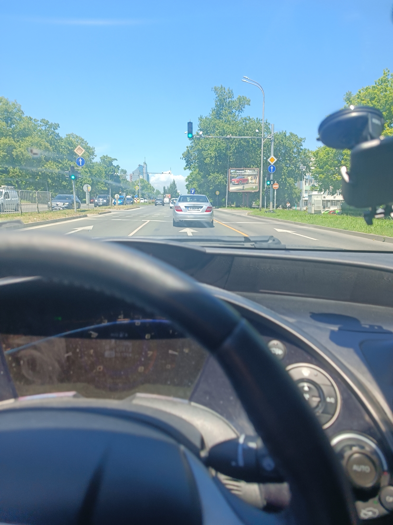 The image is taken from the driver's seat of a car driving on a sunny day in Burgas, Bulgaria. The road is a multi-lane street with trees lining the sides and modern buildings visible in the distance. There are several cars on the road, including a silver Mercedes-Benz in the foreground, a black Mercedes-Benz further up the road, and other vehicles. Traffic lights are visible and appear to be green. A large billboard advertising a car is also present. The dashboard and steering wheel of the car are in the foreground, indicating a first-person perspective of driving.