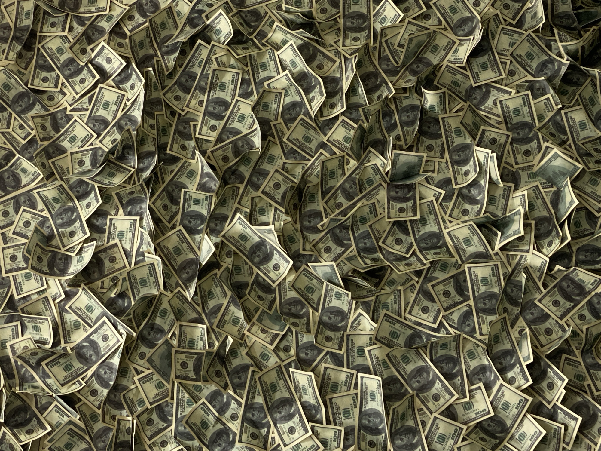 The image shows a large pile of United States one hundred dollar bills. The bills are stacked and scattered irregularly, creating a dense and overwhelming mass of money. The image does not depict any location, city, people, actions or interactions. It is purely a close-up view of the pile of money.