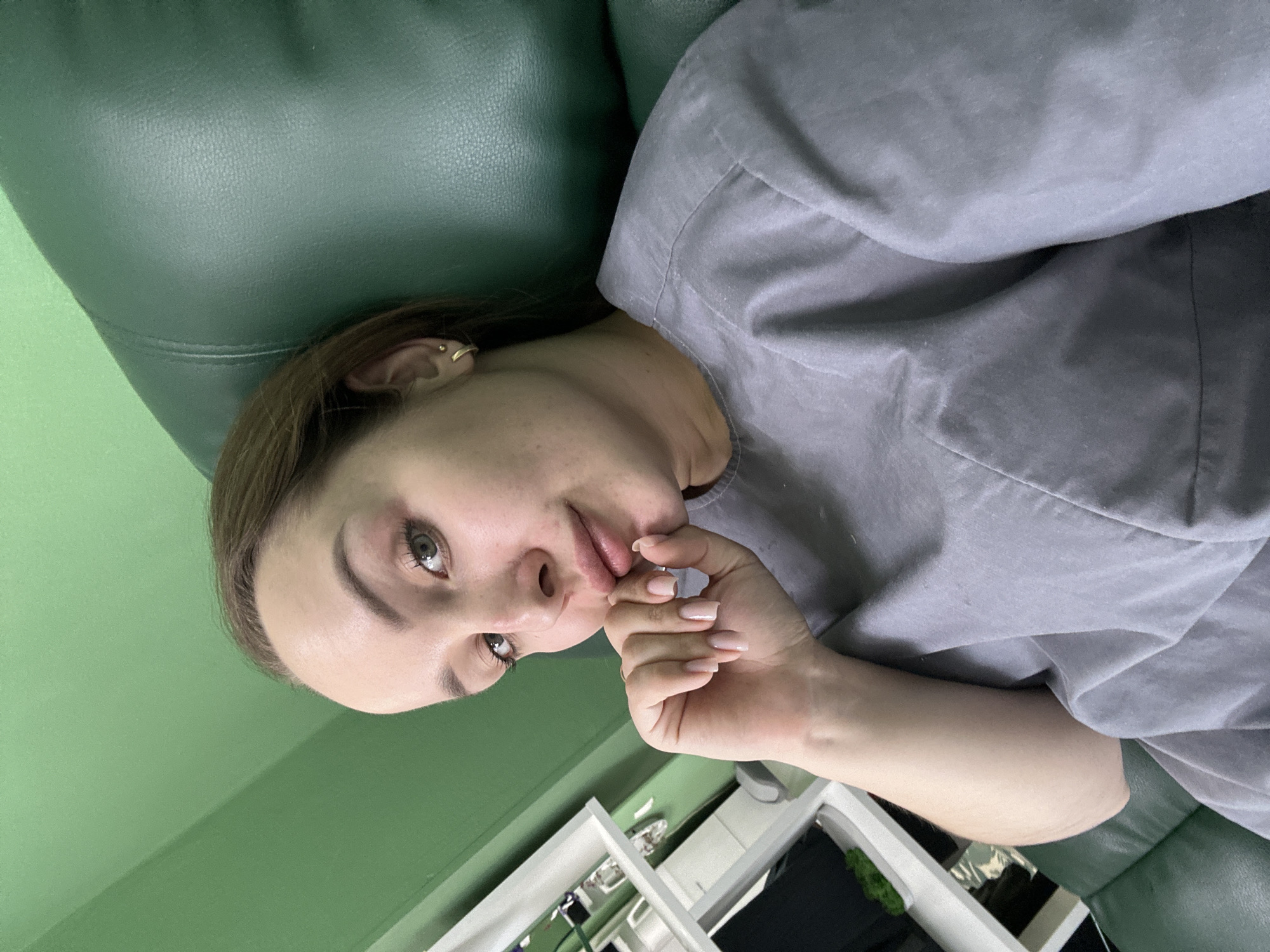 A person lies on a green leather-like sofa. The person has light skin, brown hair, and green eyes. They wear a gray t-shirt and have light pink nail polish. Their right hand is near their mouth. In the background, there is a white shelf with items on it, and a green wall is visible. The photo was taken in Chaikovsky, Russia.