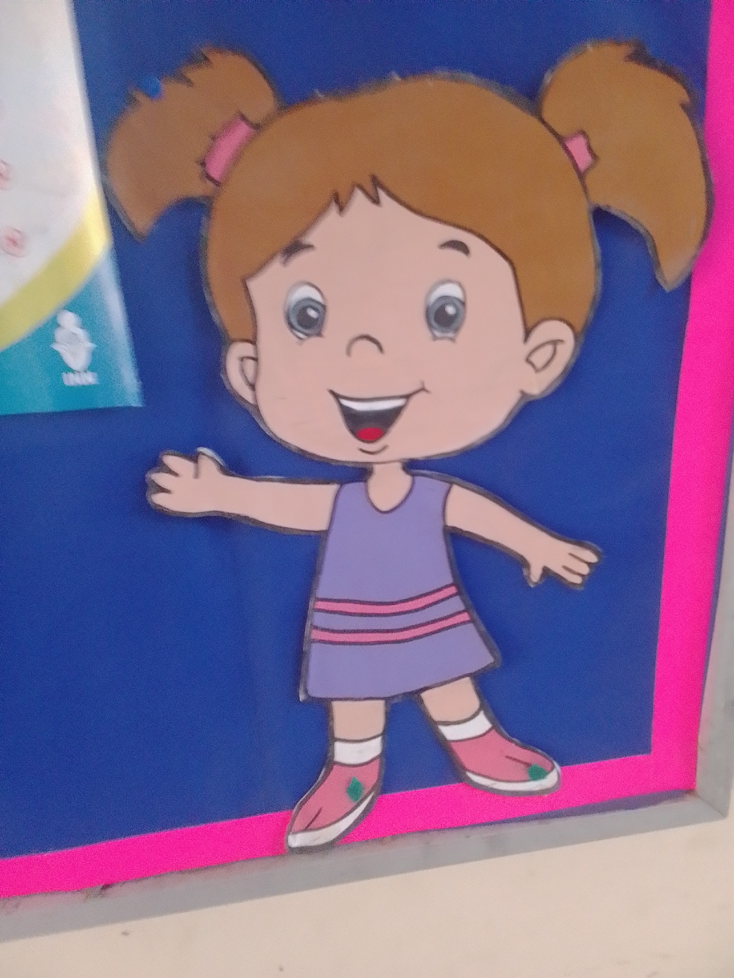 This is a close-up, eye-level shot of a colorful, cartoon-style cutout of a young girl. She has brown hair styled in pigtails with pink ribbons, and is wearing a purple dress with pink stripes. Her shoes are pink with white soles and green accents. The cutout is attached to a blue surface, possibly a bulletin board, which is bordered by a bright pink trim. To the left of the cutout, part of a white and yellow poster with some indiscernible text and a graphic of a stylized figure is visible. The overall impression is one of a cheerful and playful decoration, likely for a children's area or classroom. The lighting is bright, suggesting an indoor setting during the daytime. The visible text on the poster includes "UNIV." The location context is La Tagua, Venezuela.