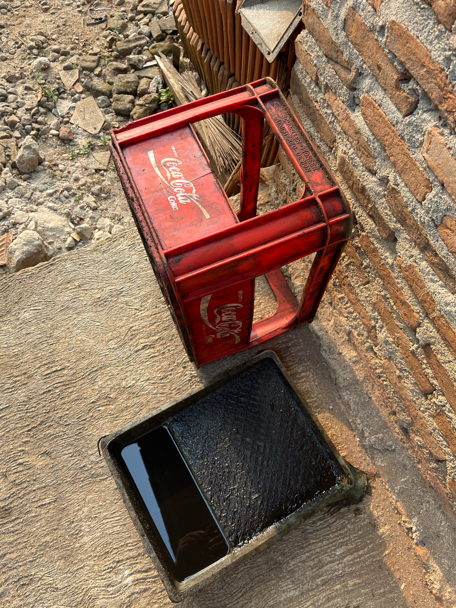 An overhead, close-up shot shows a red Coca-Cola crate placed on a textured concrete surface, next to a brick wall. The crate is oriented with its opening facing upwards, and the Coca-Cola logo is visible on its side. To the left of the crate, a black pan filled with dark liquid, possibly oil, sits on the concrete. The background consists of a pile of rubble and a stack of wooden planks. The scene appears to be outdoors, in a part of Kebumen, Indonesia.