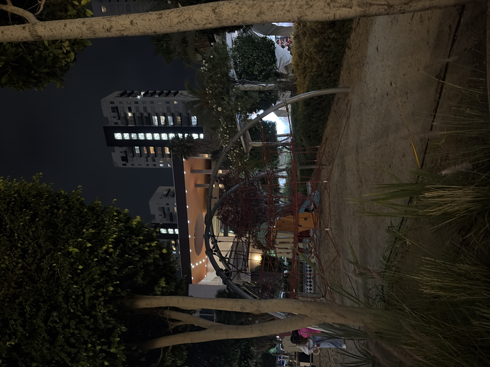 The image shows an outdoor scene at night, likely in San Bernardino Tlaxcalancingo, Mexico, given the location specification. In the background, there are buildings, including a taller one with multiple illuminated windows, suggesting residential or commercial use. There is a playground structure with a complex arrangement of red climbing elements visible in the middle ground. The foreground contains trees and vegetation, which partially obstruct the view. Some people are present at the bottom of the image, possibly interacting near the playground area. The overall illumination is low, creating a contrast between the artificial lights and the dark sky.