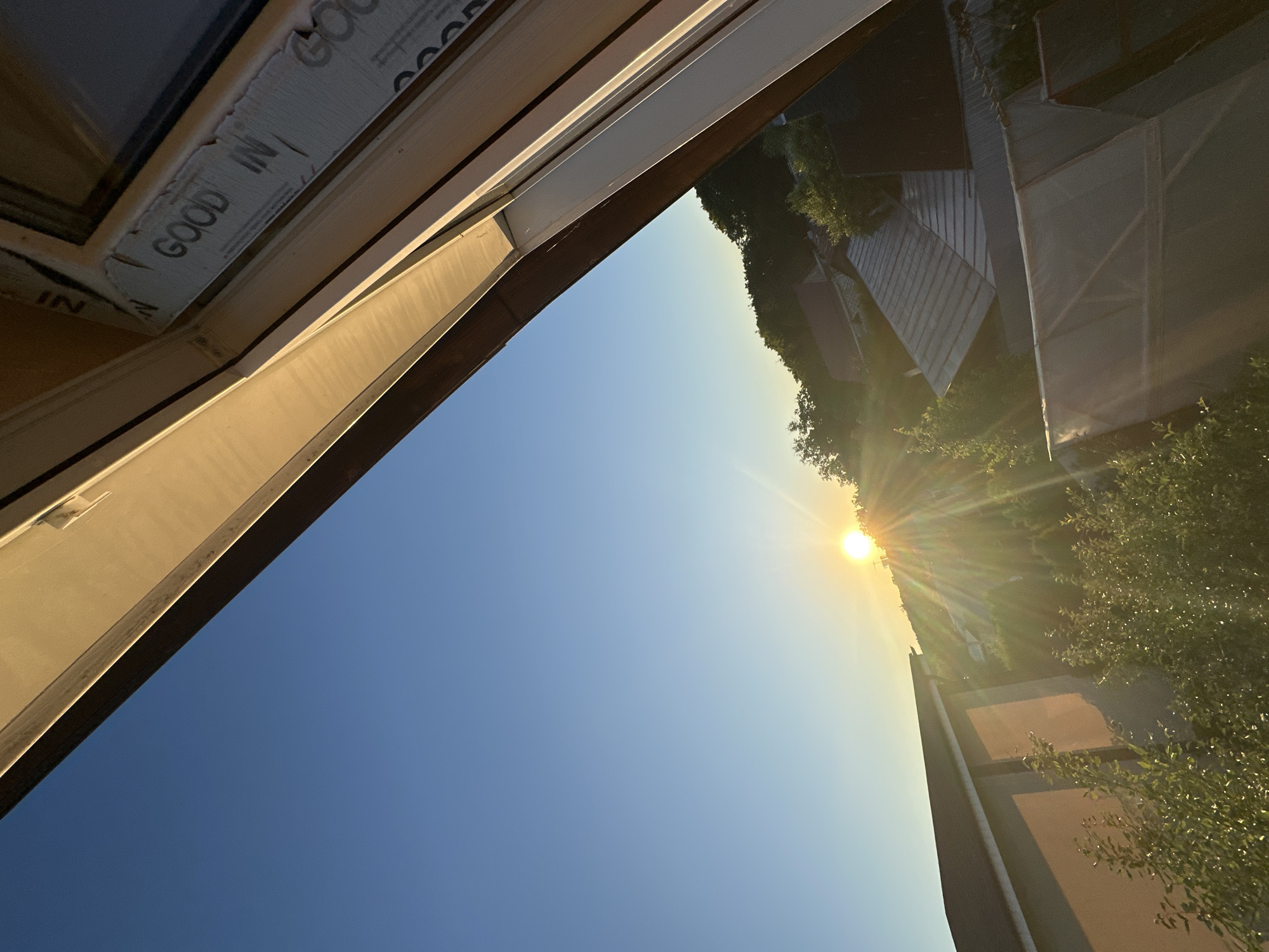 The image captures a view looking upwards through an open window frame, with the sky dominating the frame. The sun is low in the sky, casting a warm, golden light and creating lens flare effects that streak across the scene. This suggests the photograph was taken during either sunrise or sunset. The sky is a clear, vibrant blue, transitioning to a lighter hue closer to the horizon.
In the foreground, the interior of a window frame is visible. There is some protective tape with the text "GOOD IN" printed on it, indicating recent construction or installation. The window frame itself appears to be made of white or light-colored material.
Beyond the window, the scene depicts an outdoor setting. There are glimpses of trees with green foliage and some structures that appear to be part of a residential or commercial area. The angled sunlight illuminates parts of these structures, creating shadows and highlighting textures. One building in the background has a roof with a distinctive geometric pattern.
The overall mood is serene and peaceful, enhanced by the warm sunlight and clear sky. The composition, looking up and outwards, evokes a sense of openness and possibility. The specific location is identified as Nizhny Novgorod, Russia, which adds a geographical context to the captured moment. No people are visible in the image.
