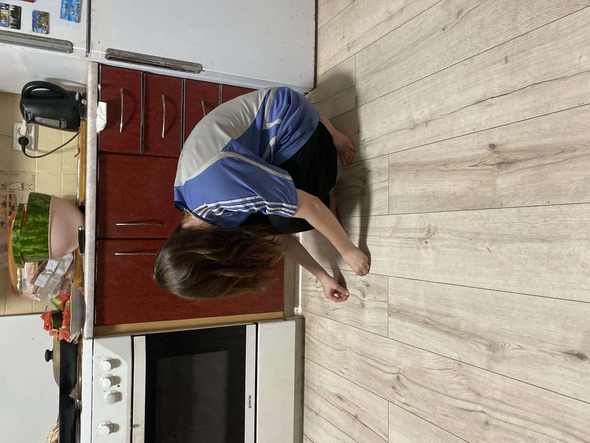 A person crouches on a light wood laminate floor in a kitchen. The person is wearing a blue and gray athletic shirt with white stripes on the sleeves and black pants. They are facing away from the camera and appear to be picking something up off the floor. The kitchen has white appliances, including a stove, and red cabinets. A black electric kettle is plugged into the wall above the counter. Sliced watermelon is visible on a plate on the counter. The location is Chornomorsk, Ukraine.