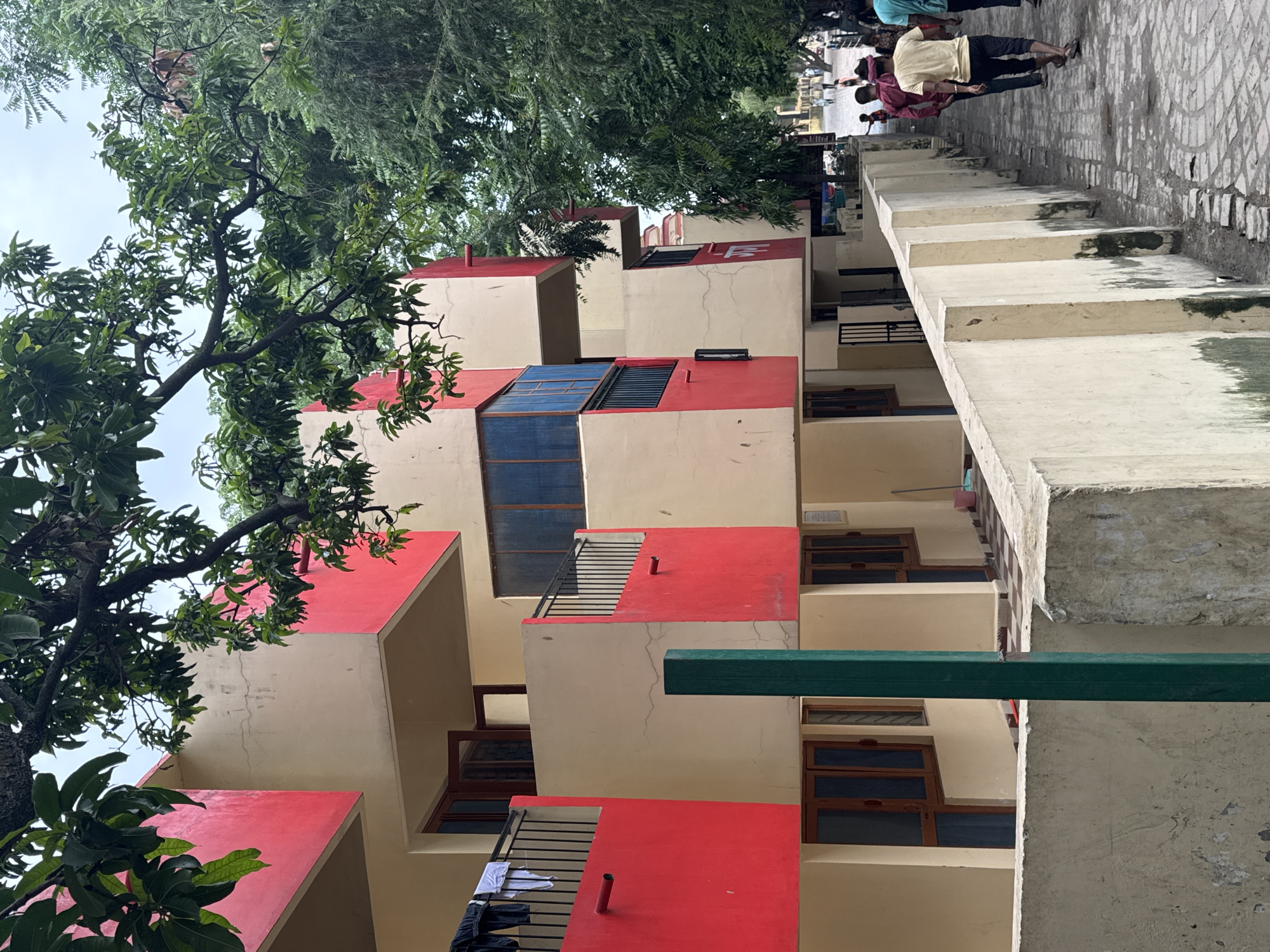 The image depicts an exterior view of a building with a distinctive architectural style, likely a residential complex or guesthouse, located in Rishikesh, India. The building features a color scheme of cream and bright red, with prominent red sections accentuating balconies and facade elements. Dark-framed windows and balconies with black railings are visible. Several open windows with wooden frames and glass panes are present, with laundry hanging on a clothesline outside one of the balconies. A tall, green vertical post stands in the foreground, adjacent to a low, concrete wall.
To the right of the building, a tree with lush green foliage casts shadows, and beyond that, a paved walkway runs alongside the building. Several individuals, including men and children, are walking on the path. One man in the foreground is wearing a yellow t-shirt and dark pants, while another in a red shirt is ahead of him. The walkway is made of interlocking bricks and is bordered by a low, concrete barrier. In the distance, more trees and structures are visible, suggesting a developed urban or semi-urban area. The overall atmosphere suggests a warm, possibly humid, day. The presence of people in traditional Indian attire and the architectural style suggest the location is indeed in India, consistent with Rishikesh.